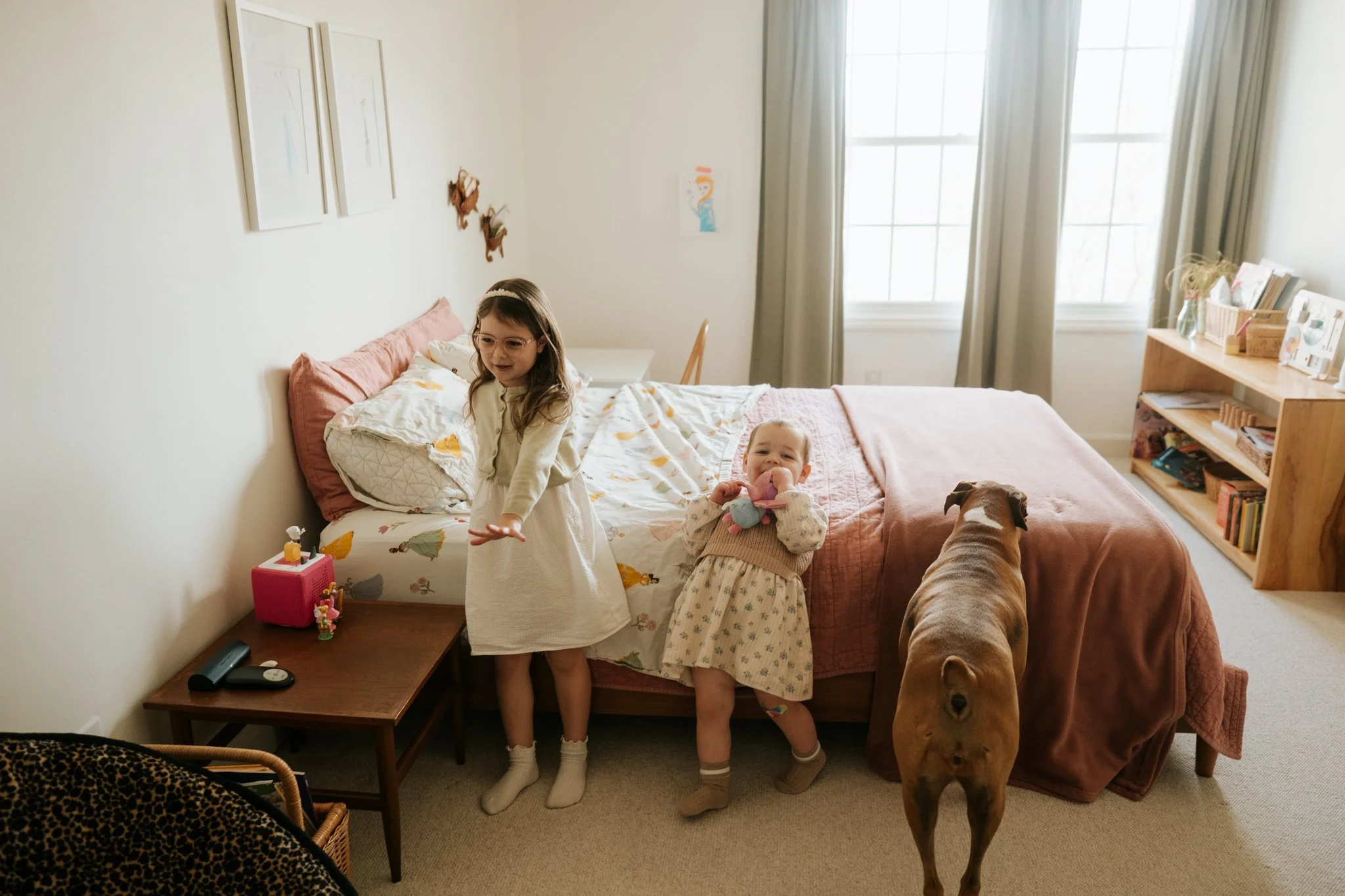 Two young girls and a dog in a domestic bedroom. One girl stands on a bedside table, the other, holding a stuffed animal, stands on the floor. The dog is a large brown one, standing near the bed, looking towards the girls. The room has a window with 
