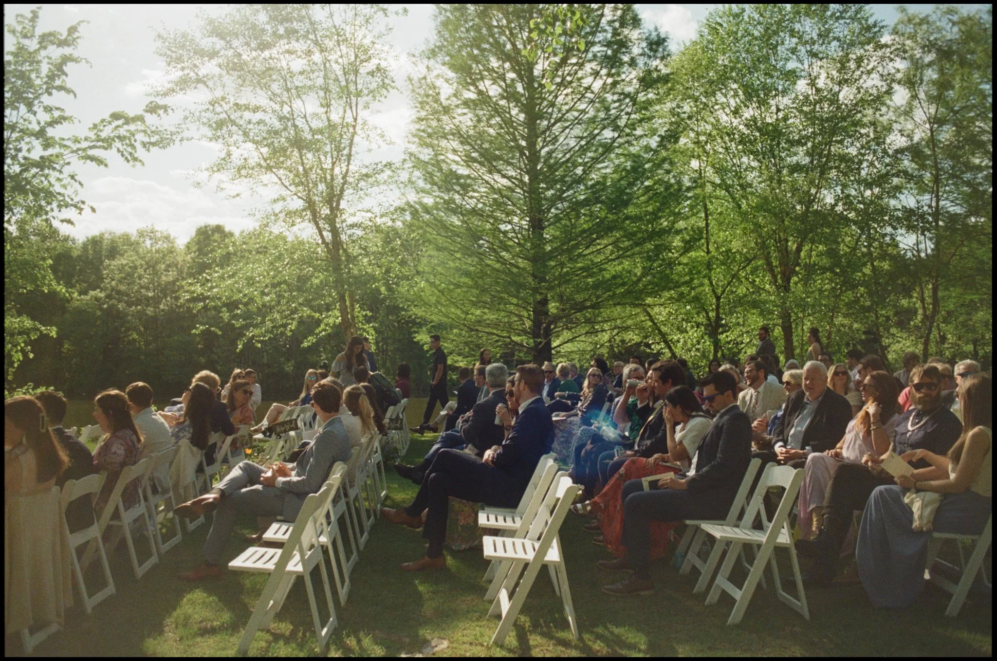 People attending an outdoor event seated on white chairs in a green park with tall trees and sunlight filtering through the leaves.
