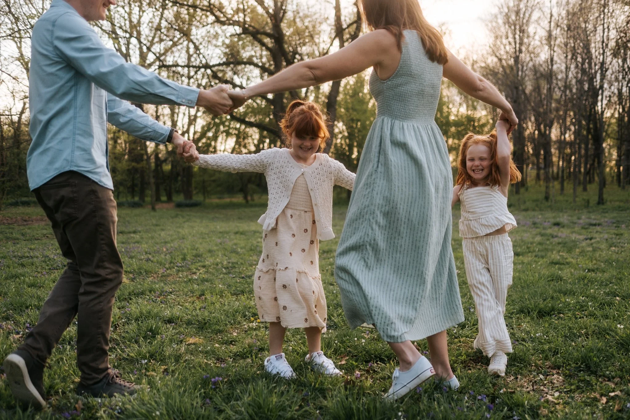 A family of five holding hands in a circle outdoors on a grassy field with trees in the background during sunset, smiling and enjoying each other's company.