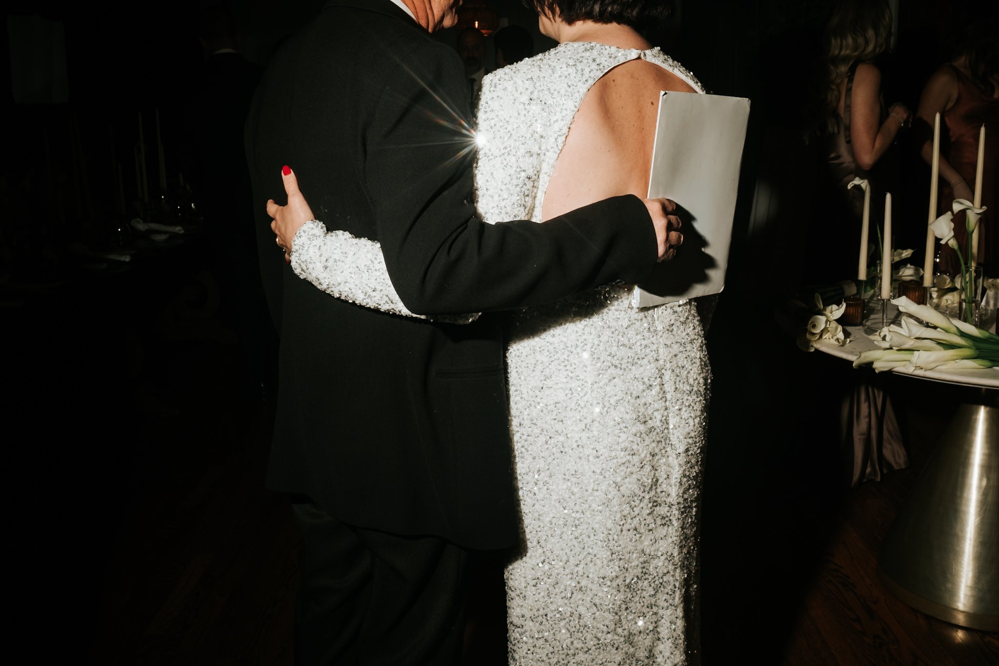 A couple dancing at a formal event. The woman is wearing a sparkling, sleeveless dress, and the man is dressed in a dark suit. The setting appears to be a dimly lit banquet hall with floral arrangements.