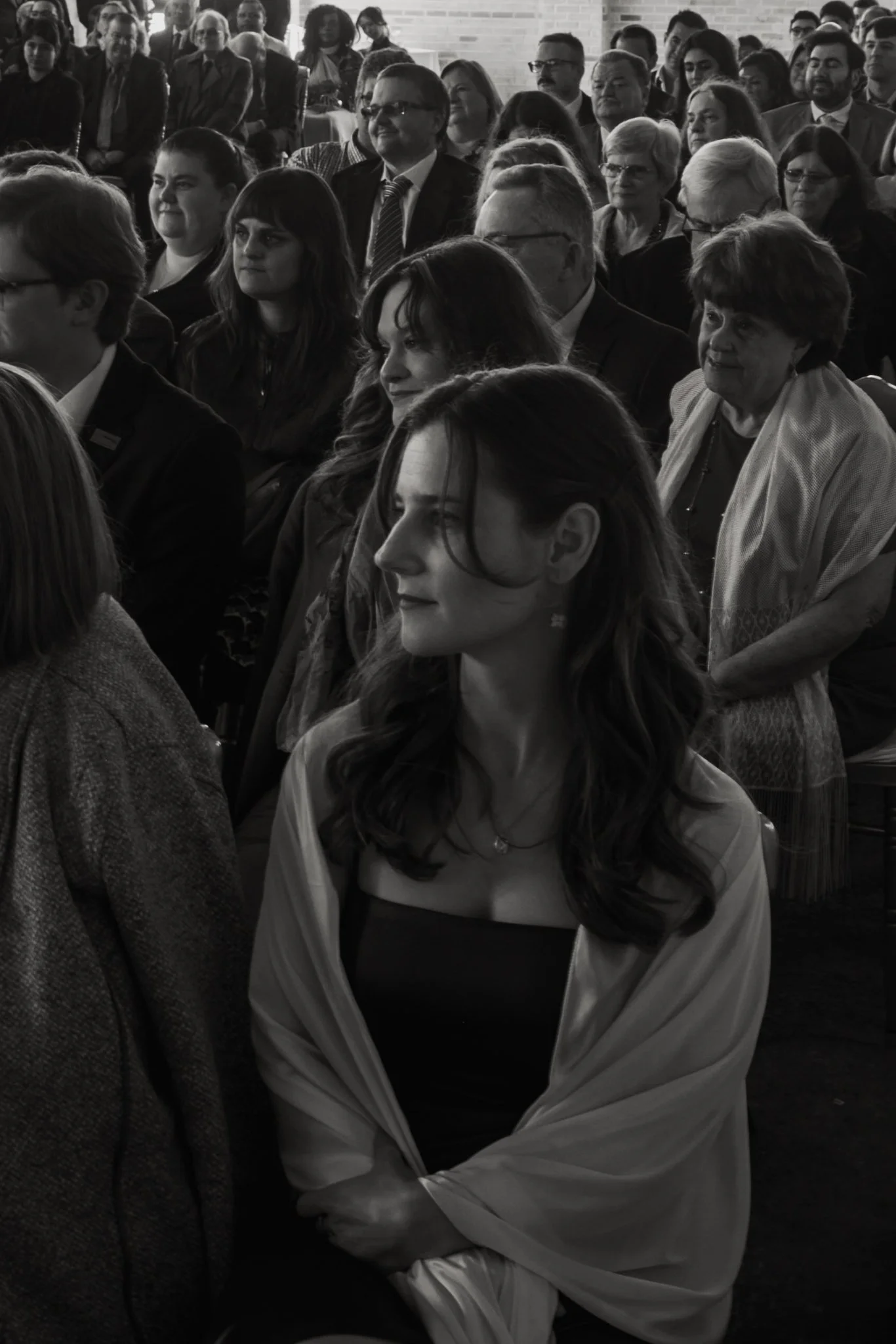 Black and white photo of a large gathering of people seated, attending an event, with some looking forward and others engaged in conversation.