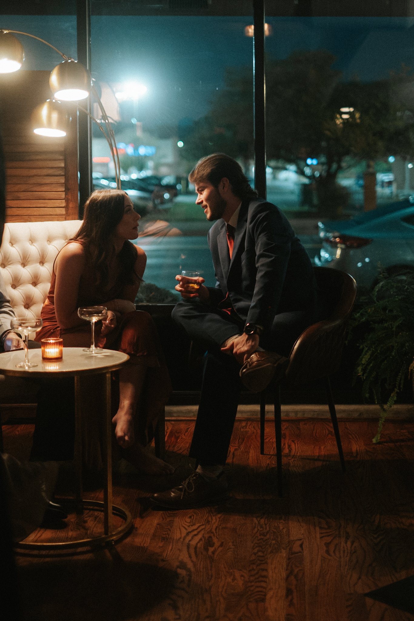 A man and woman having a conversation at a table in a restaurant or bar during the evening, with drinks in their hands and warm lighting.