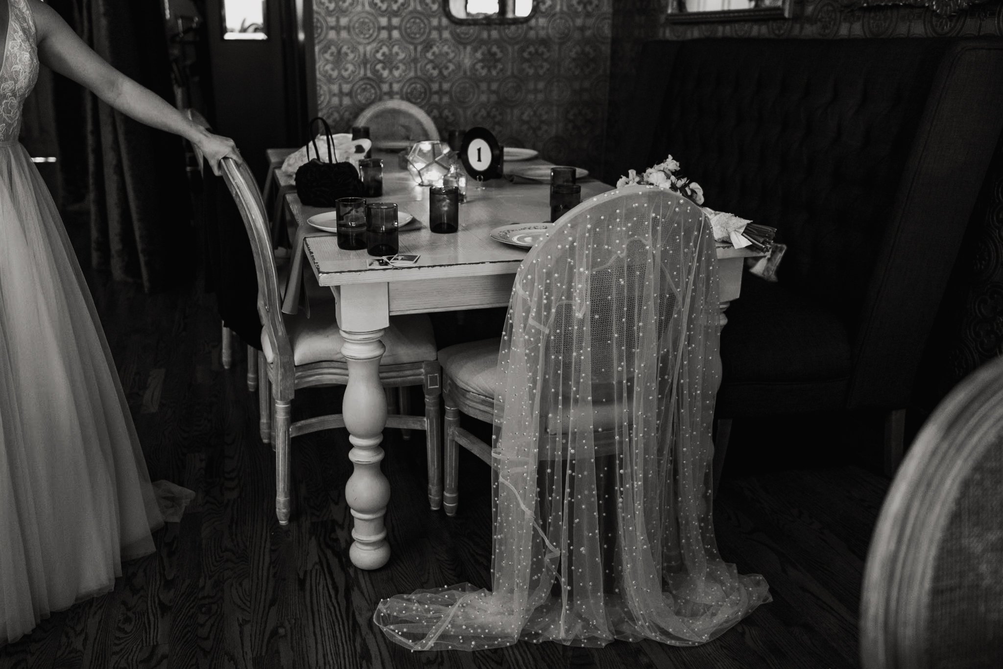 A dining table set with plates, glasses, and a centerpiece, surrounded by wooden chairs with sheer, beaded chair covers, in a decorated room with patterned walls.