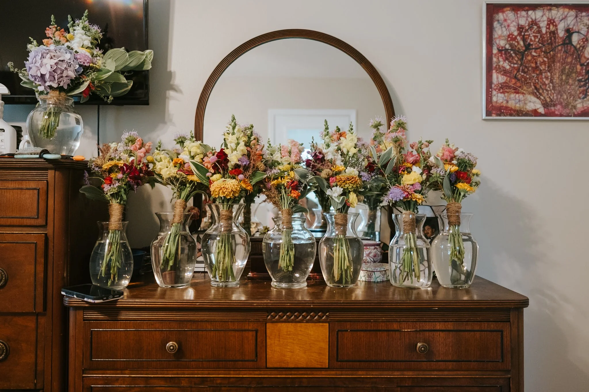 Multiple glass vases with colorful flower arrangements on a wooden sideboard in front of a mirror, with additional floral arrangements and wall art in the background.