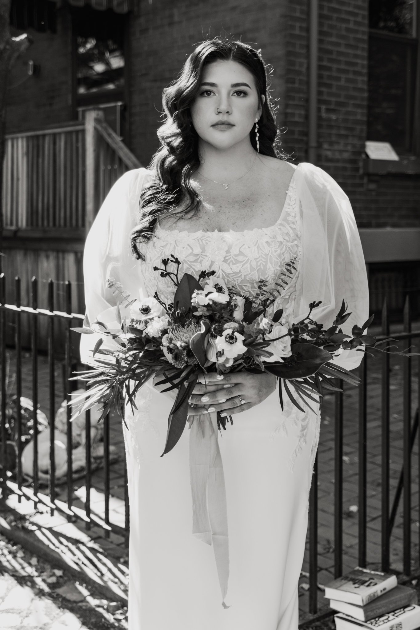A woman with dark wavy hair, dressed in a white gown, holding a bouquet of flowers, standing outdoors near a brick building and a metal fence, with books on the ground nearby.