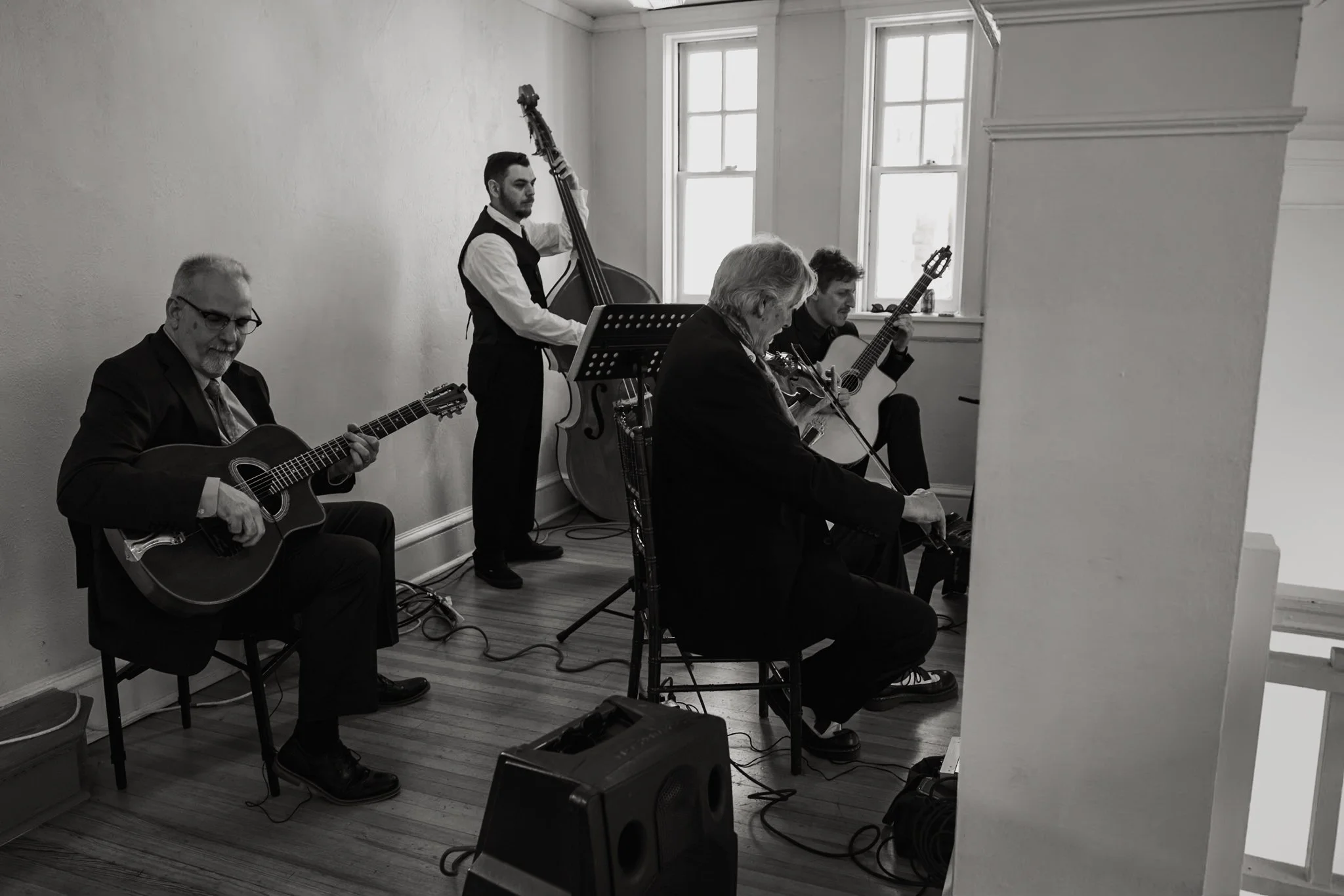 Four men playing guitars and a man playing a double bass, performing music in a room with white walls and large windows.