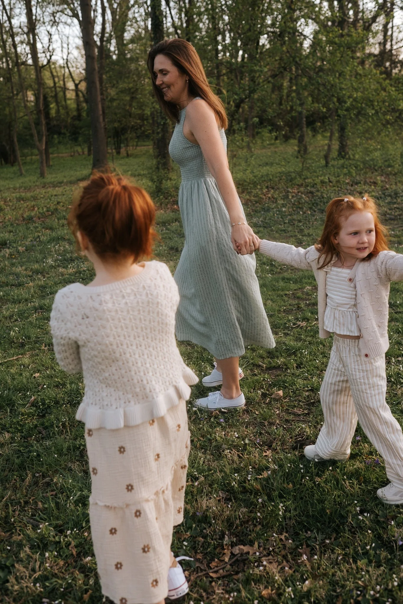 A woman in a gray dress holding hands with two children with red hair in a grassy park with trees, during late afternoon or early evening.