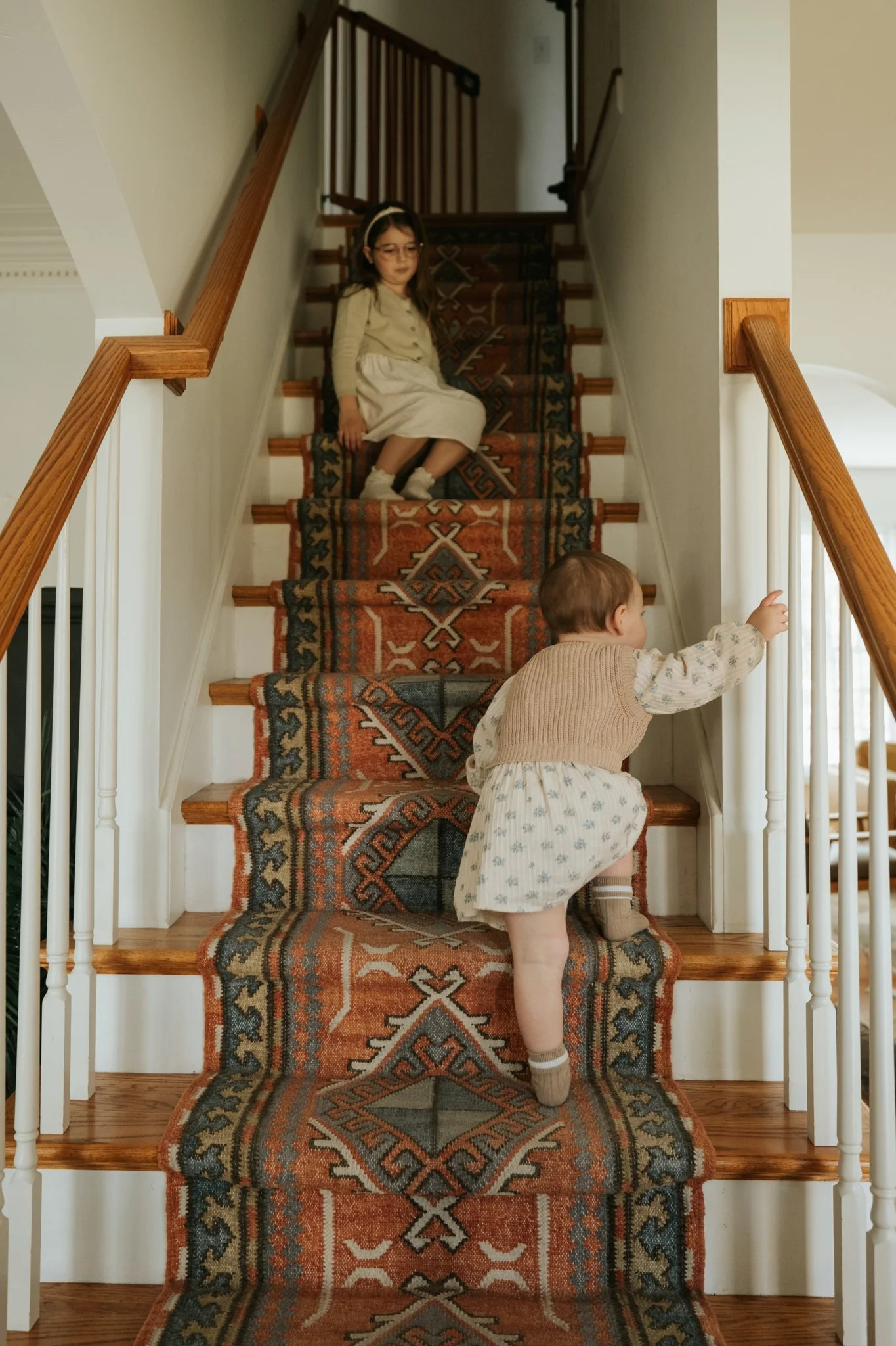 A young girl with glasses and a headband sits on a staircase with a patterned runner rug, while a toddler in a light sweater and skirt explores near the bottom of the stairs.
