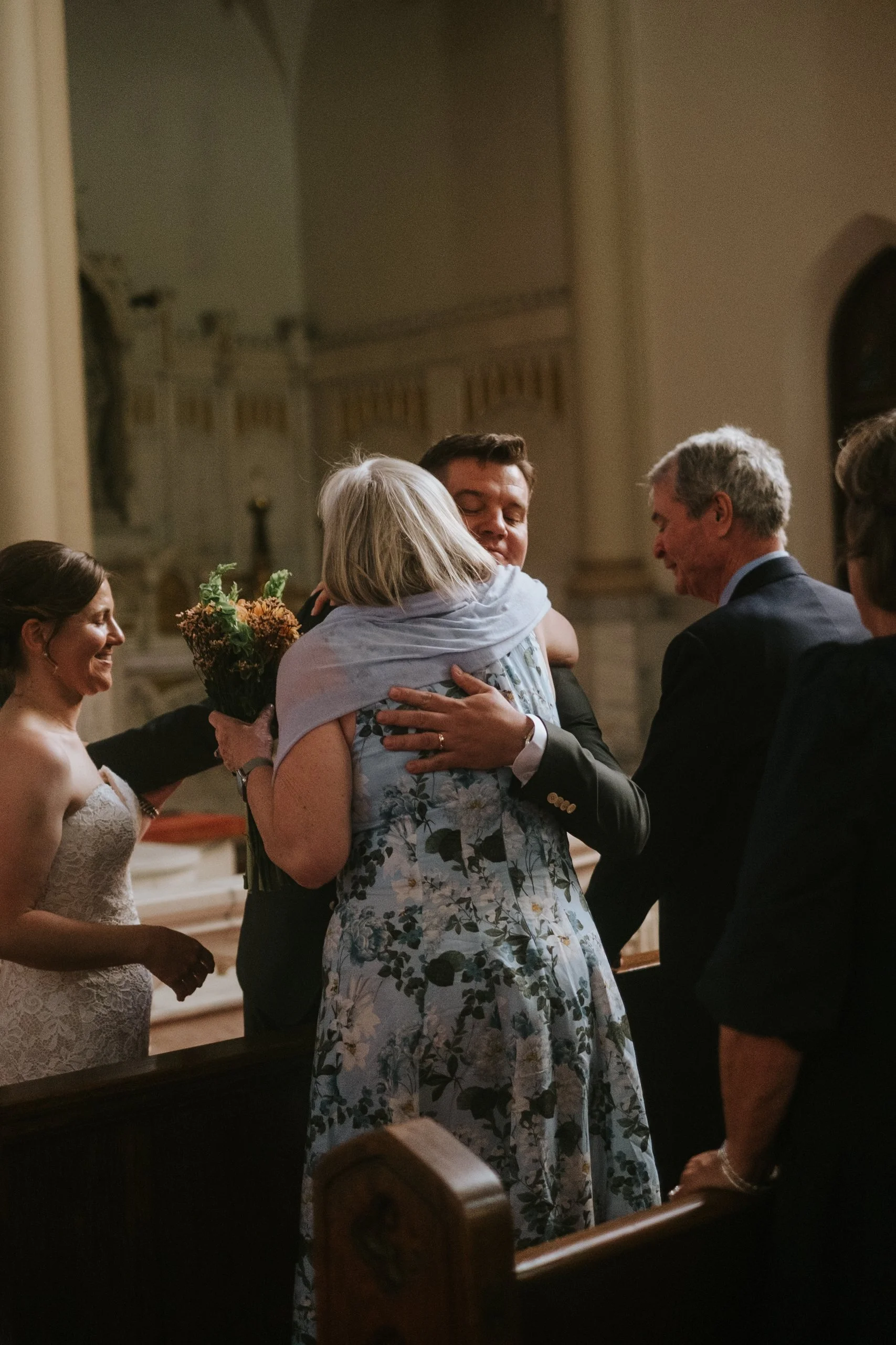 People in formal attire hugging and smiling during a wedding ceremony in a church or chapel.