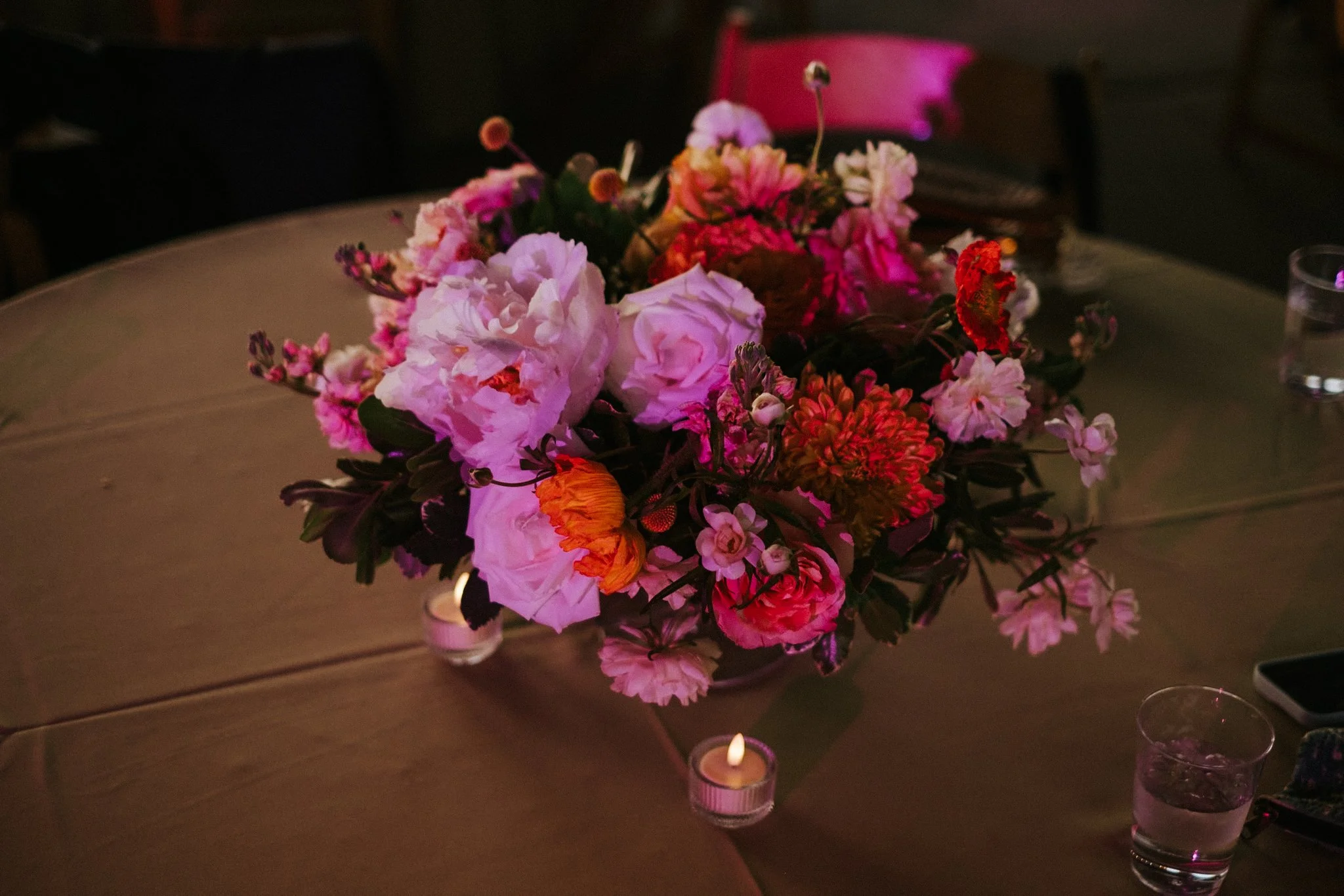 A centerpiece of pink, white, and red flowers on a table, illuminated by small candles in glass holders.