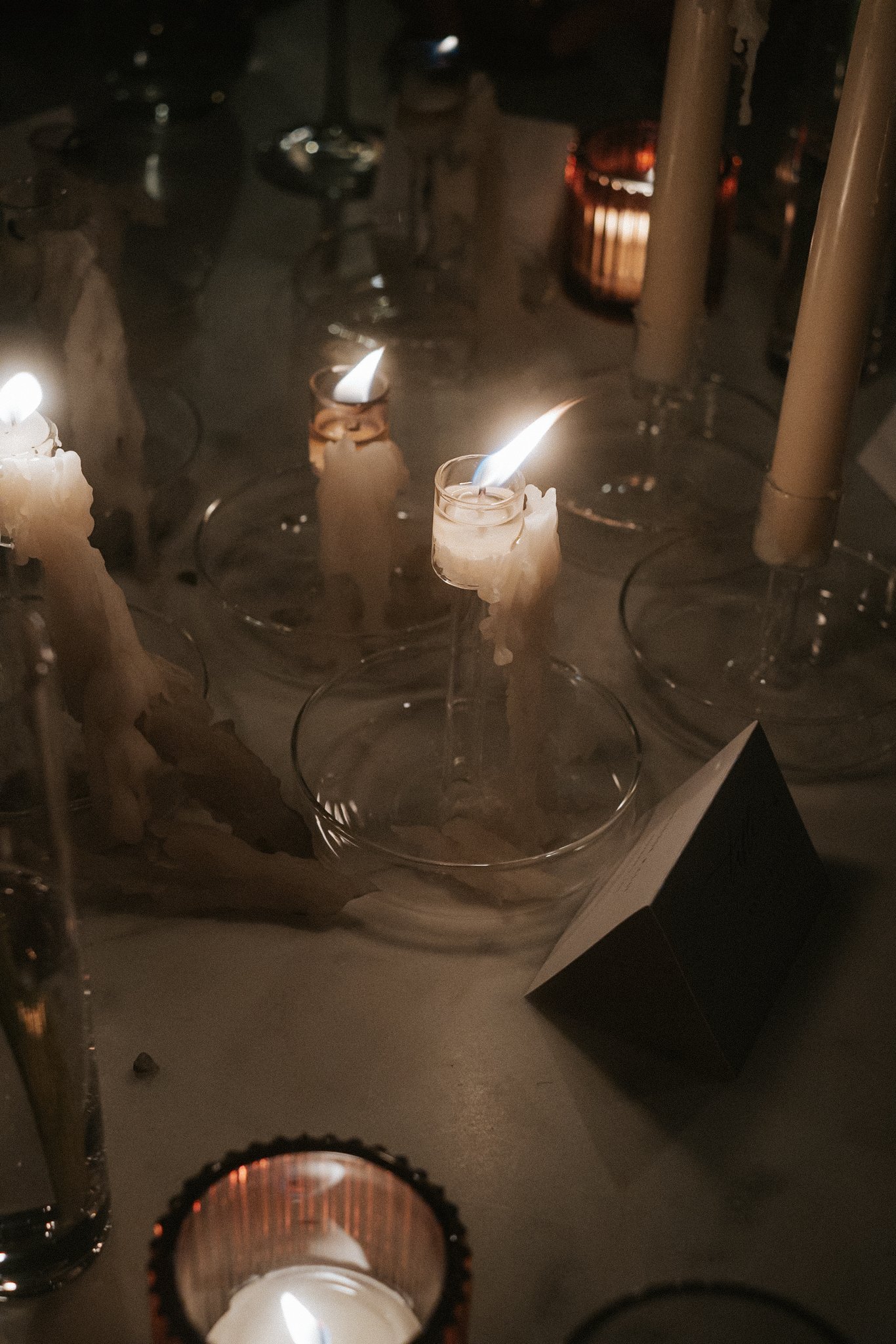 Several lit candles in glass holders on a surface with some reflecting in the background. The overall scene is dimly lit and cozy.