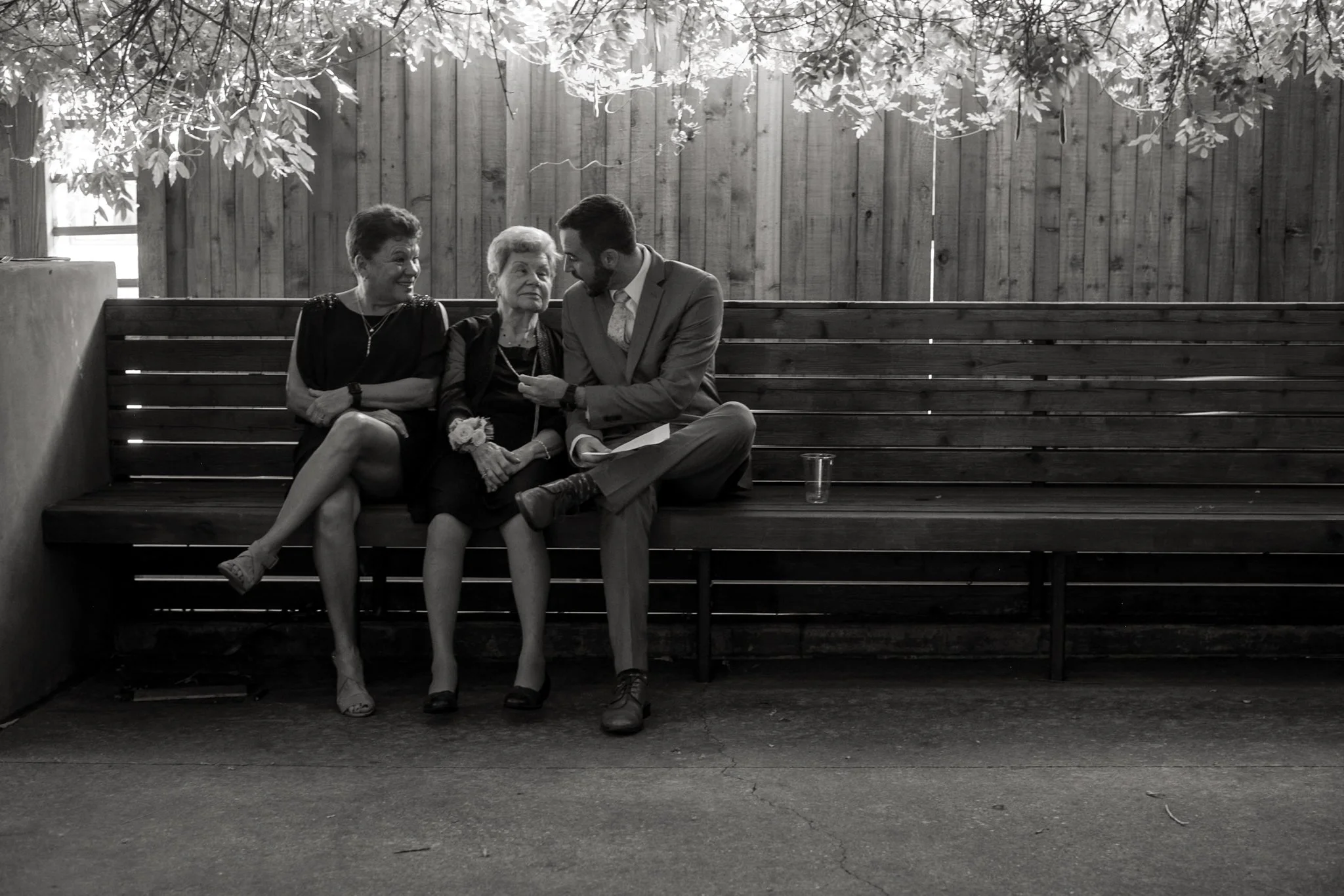 Black and white photo of three people sitting on a wooden bench outdoors, engaged in a conversation. An elderly woman in the middle is being flirted with by a young man on her right, while a woman on her left looks on smiling. There are trees and a w