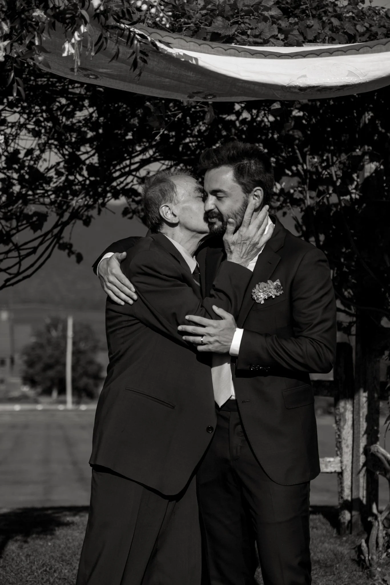 An elderly man and a younger man in formal suits are kissing and embracing outdoors. Trees and a street are visible in the background.