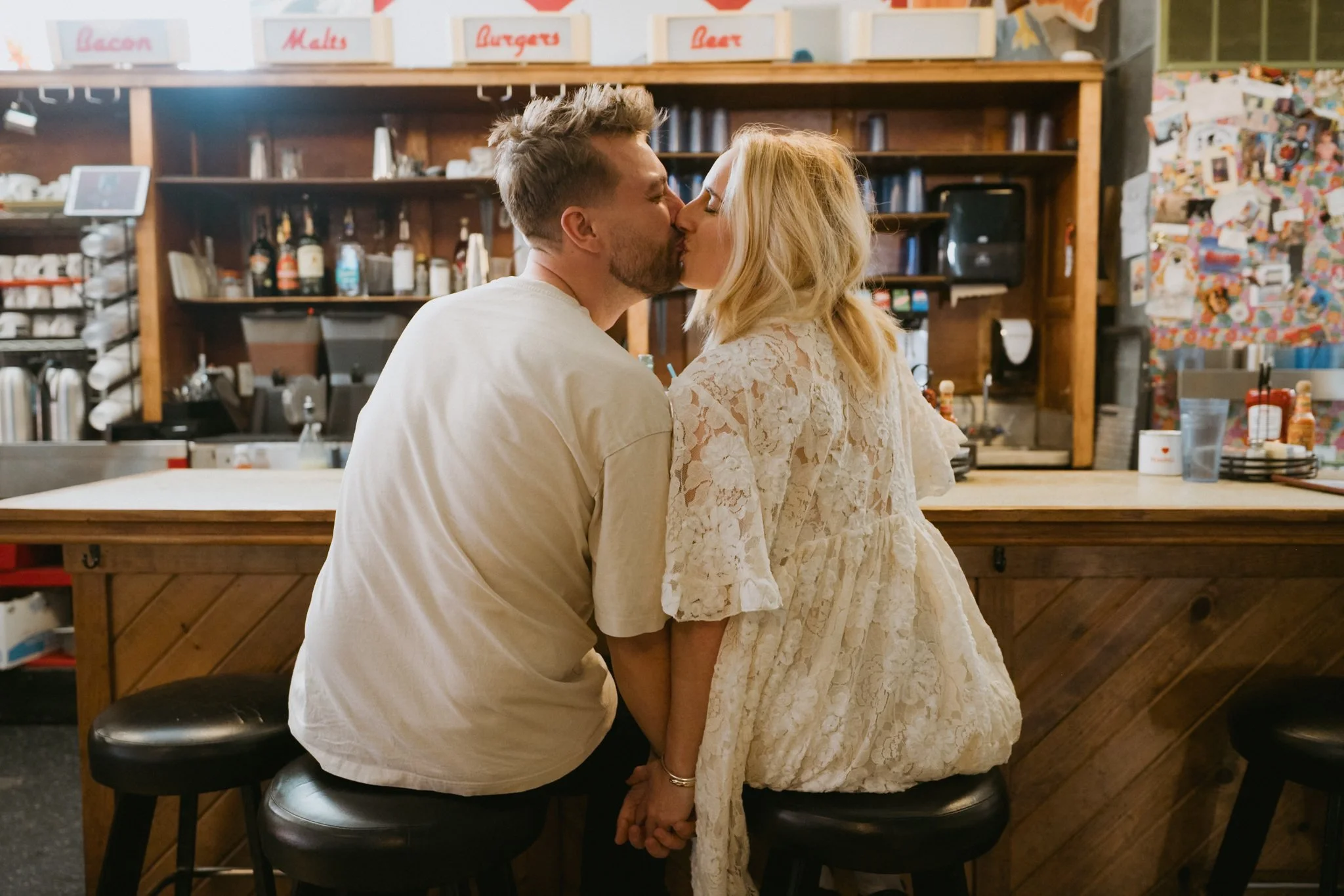 A couple kissing in a cafe, seated with their hands clasped, the man wearing a white t-shirt and the woman in a lace dress.