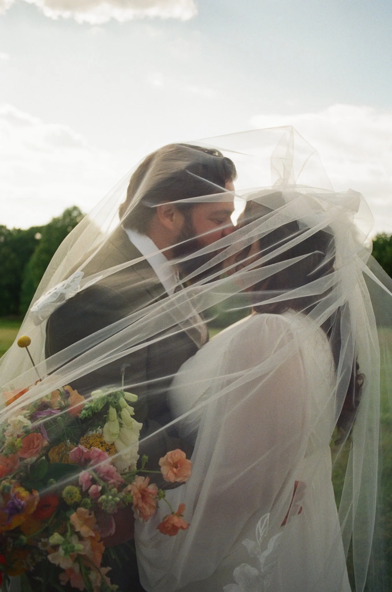 A man in a suit and a woman in a white dress embrace and kiss under a sheer white veil, holding a bouquet of flowers outdoors with a cloudy sky and green trees in the background.