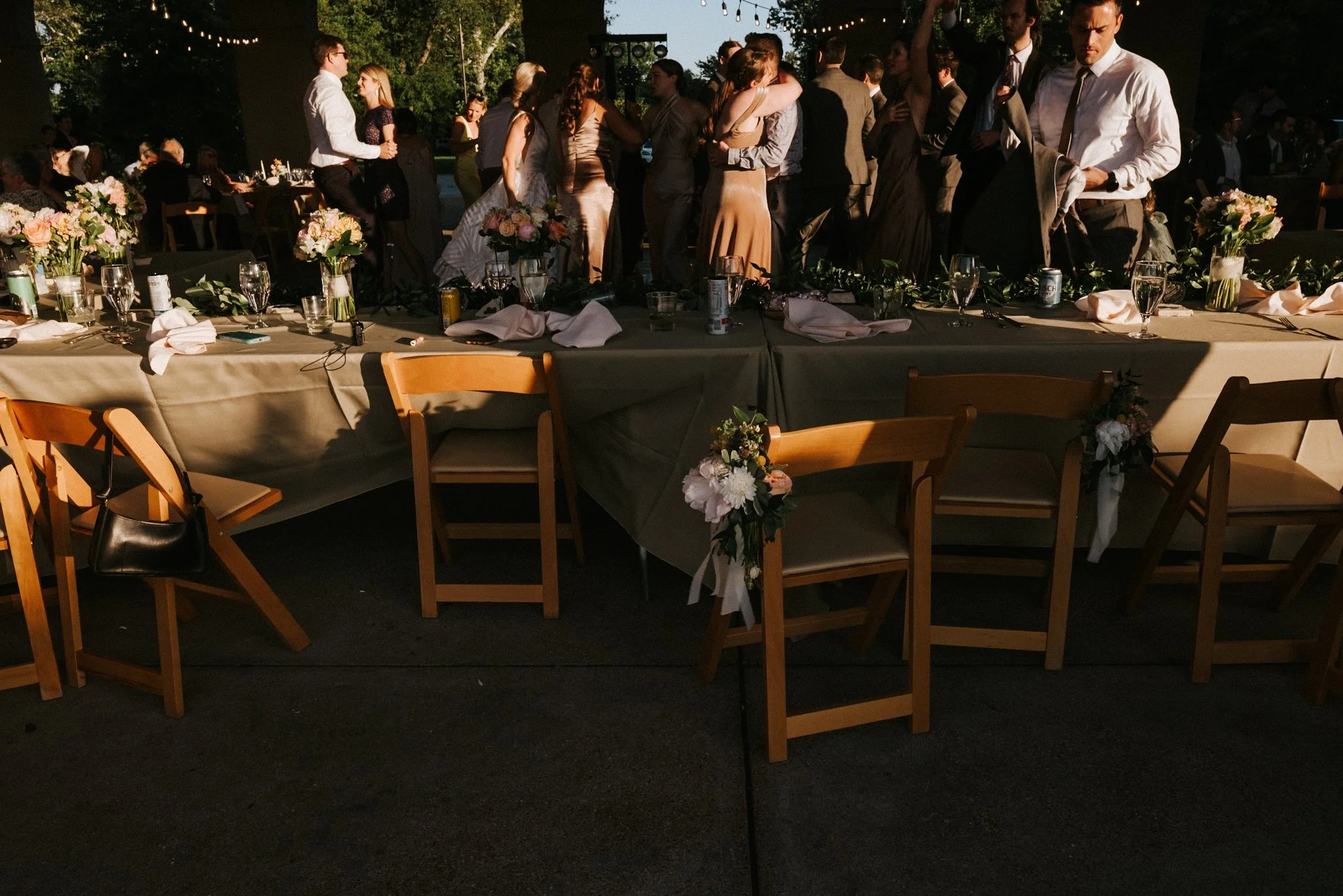 Wedding reception with guests dancing outdoors in the evening, decorated tables with floral centerpieces, and string lights overhead.