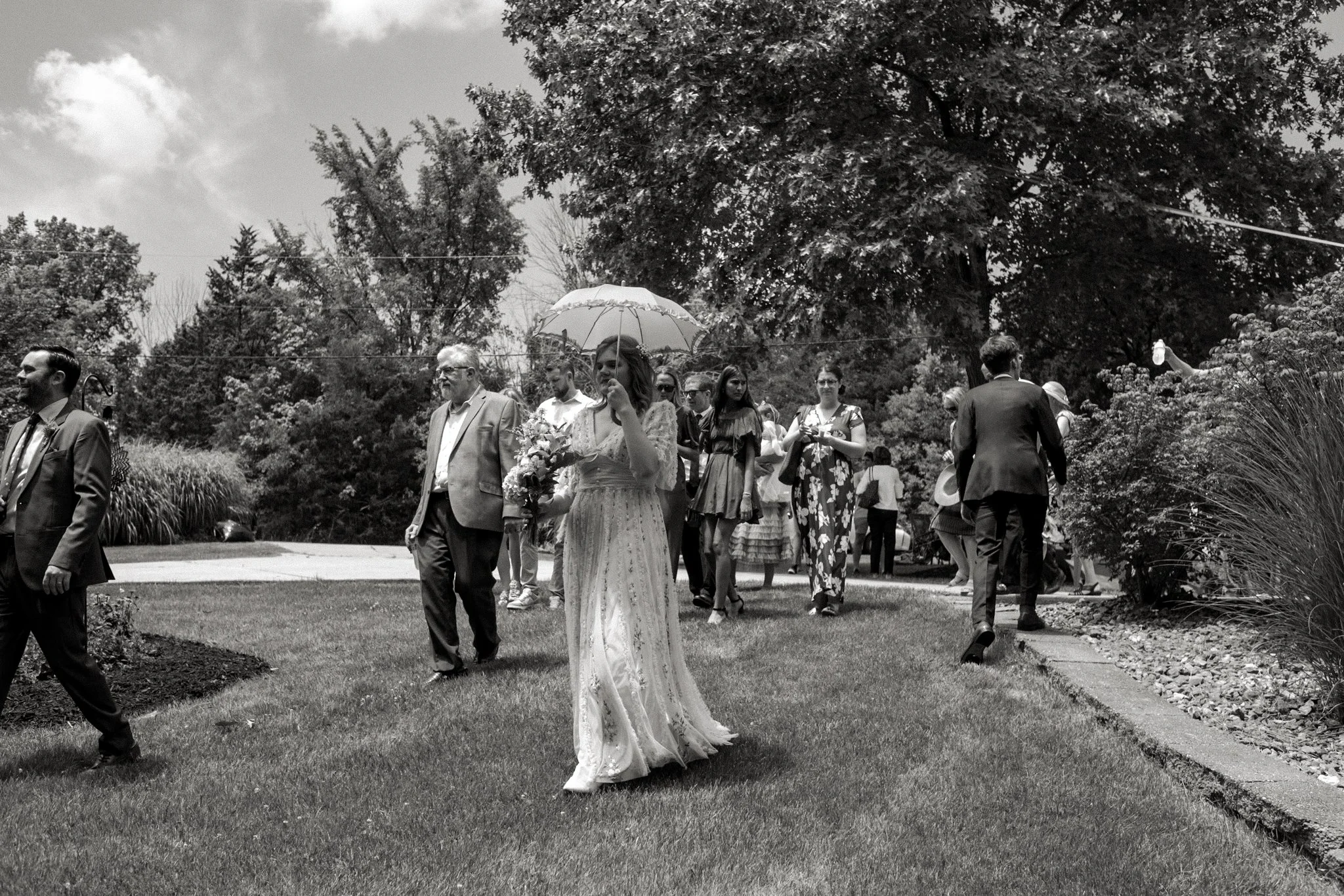 Black and white photo of a wedding procession with a bride holding a bouquet and walking outdoors on a sunny day, surrounded by guests and lush trees.