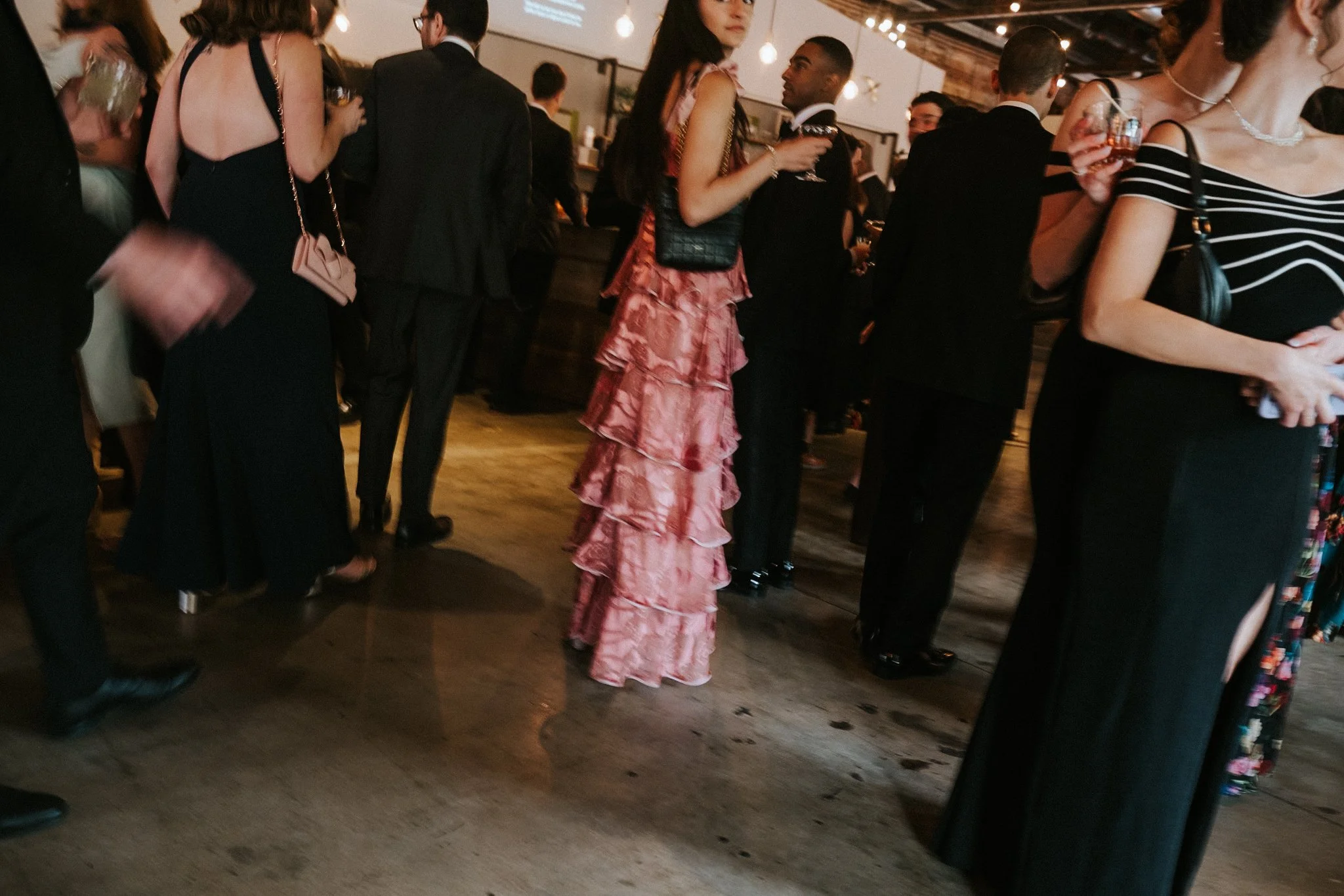 A crowd of people dressed in formal and semi-formal attire, holding drinks, at a social gathering or party in a warmly lit indoor setting.