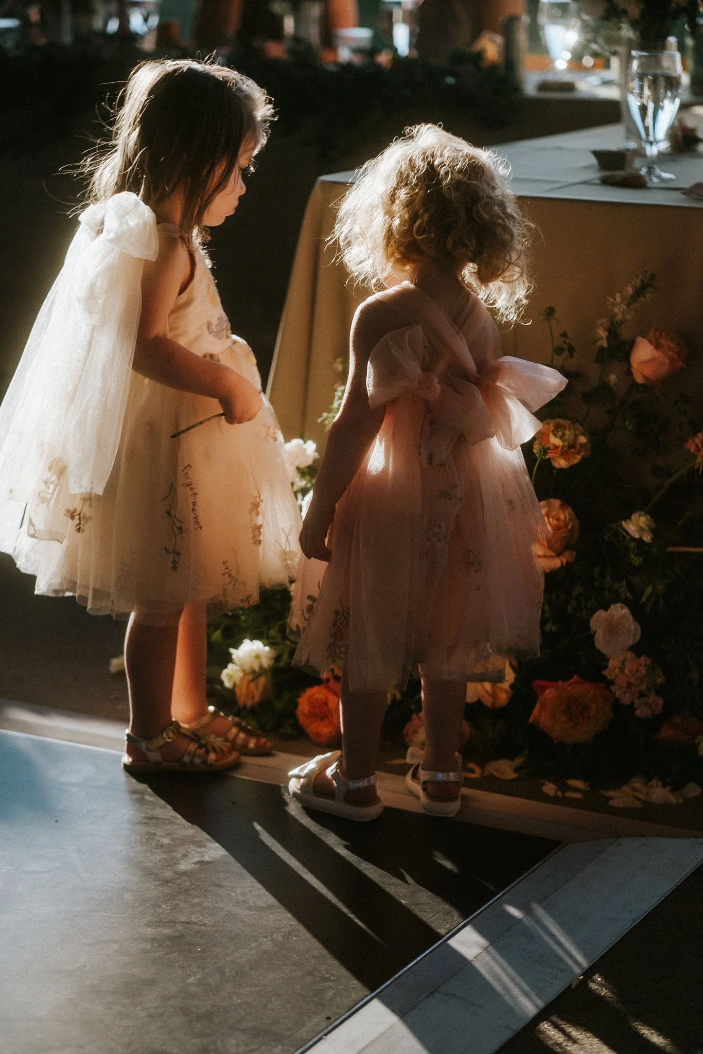 Three young girls in pastel dresses and sandals look at flowers on a stage during a performance, with sunlight illuminating their hair and the scene in a warm glow.