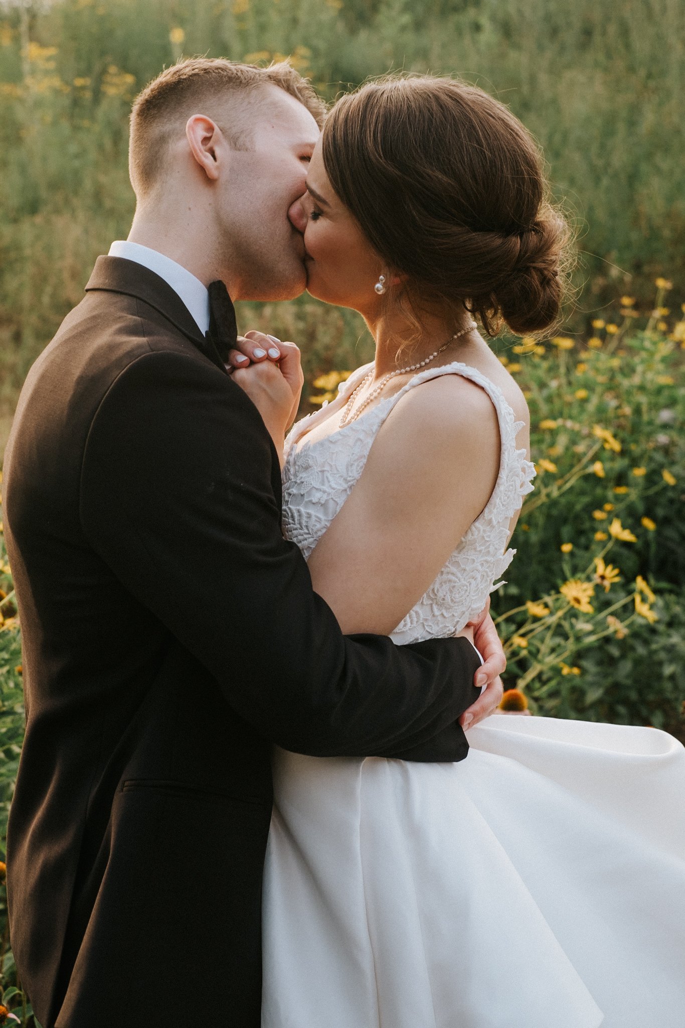 A bride and groom sharing a kiss outdoors, with the groom in a black tuxedo and the bride in a white wedding dress, surrounded by flowers and greenery.