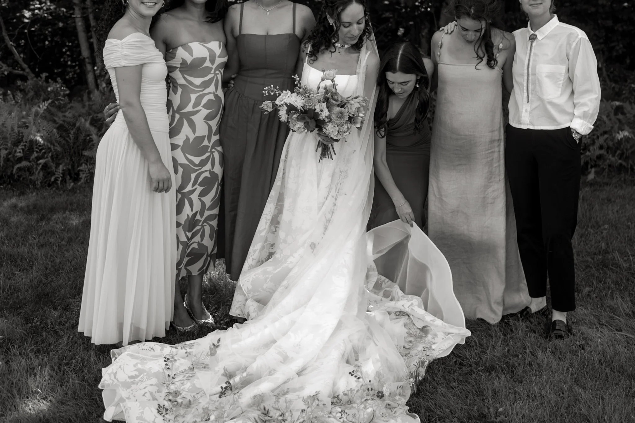 Group of women and one man standing outdoors, with some women dressed in evening gowns and holding a bouquet of flowers, likely at a wedding or special event.