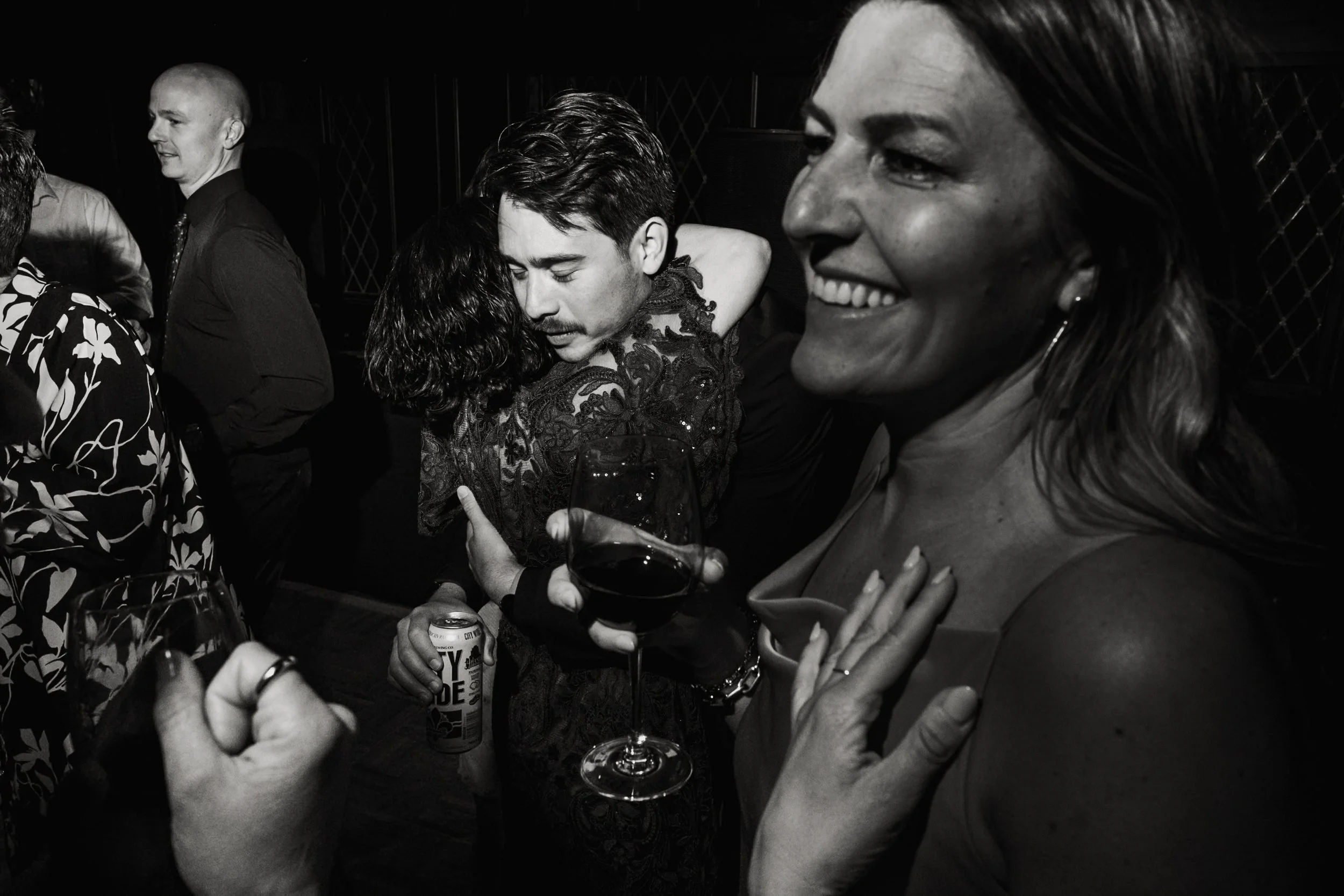 People at a party, two women hugging, holding wine glasses, smiling, with one woman wearing a floral dress, the other with her hair down, in a black and white photo.