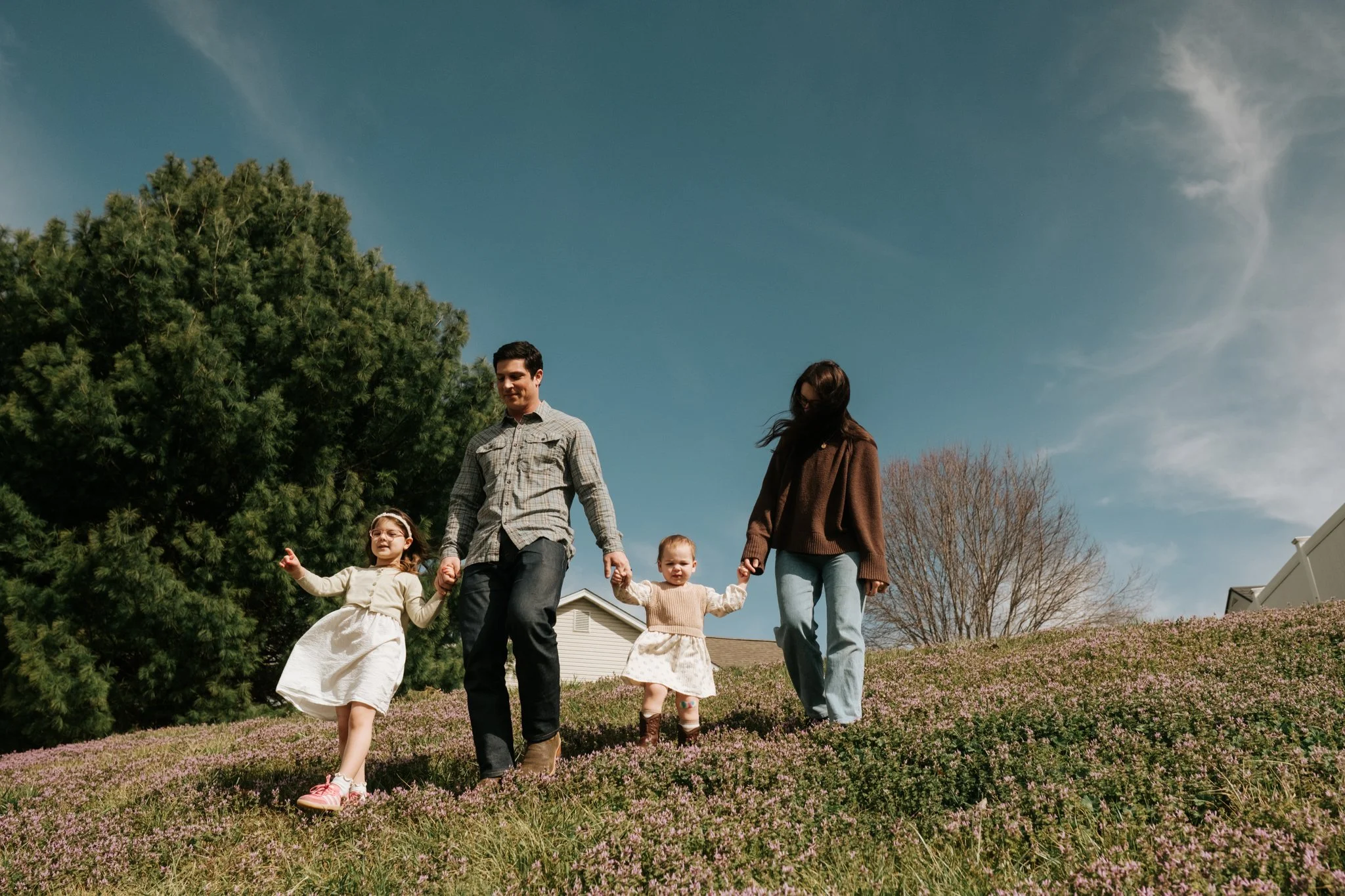 A family of five walking outdoors on a grassy hill with purple flowers, holding hands, with trees and a house in the background under a partly cloudy sky.
