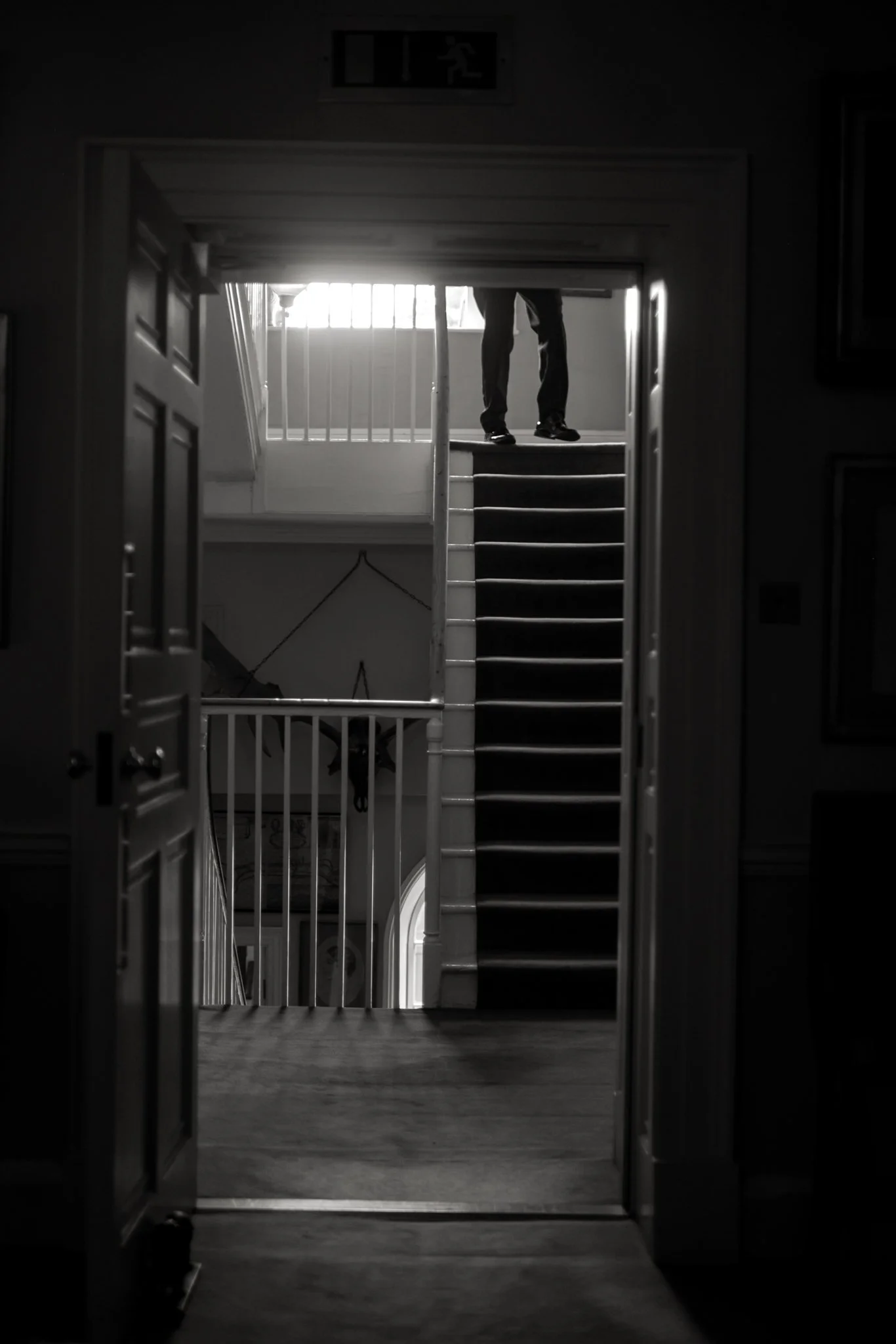Looking through a doorway into a dimly lit room with a staircase and a person standing at the top, partially visible from the waist down, with natural light coming from a window.