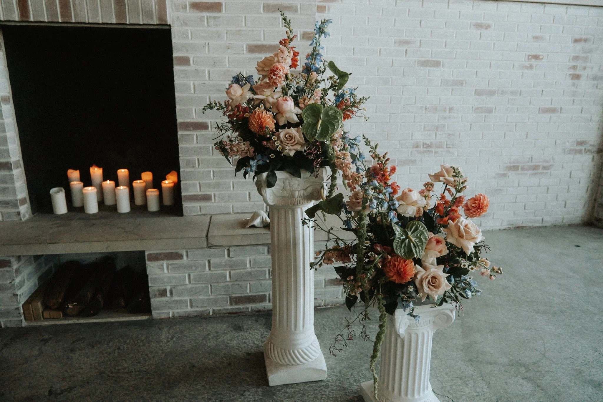 A tall white urn filled with a large, elaborate bouquet of pink, peach, blue, and white flowers, placed on a white pedestal in front of a white brick fireplace with lit candles inside.