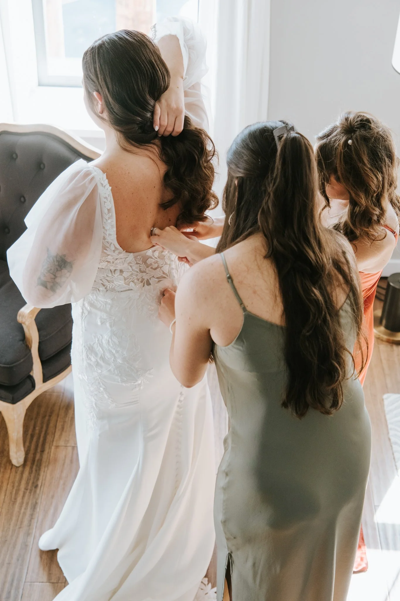 A woman in a white dress with floral embroidery on the back is standing while two women in casual dresses assist her with her wedding gown. They are indoors near a window with natural light.