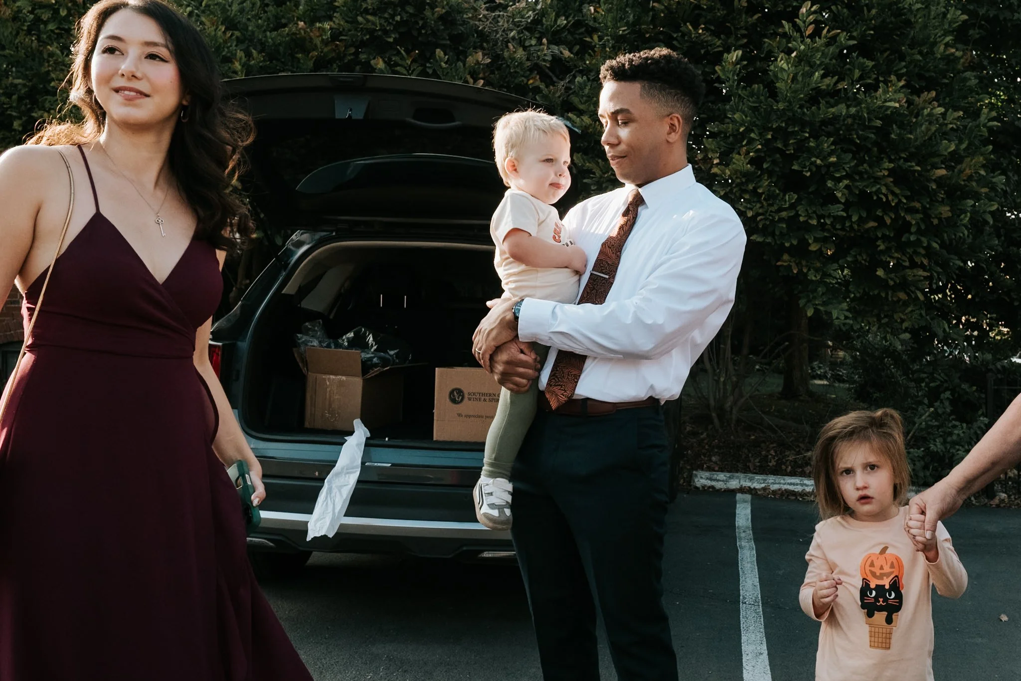 A young woman in a burgundy dress standing outdoors, a man holding a small girl in a white shirt, while a girl with a Halloween pumpkin shirt looks at the camera, with an open trunk of a car and green bushes in the background.