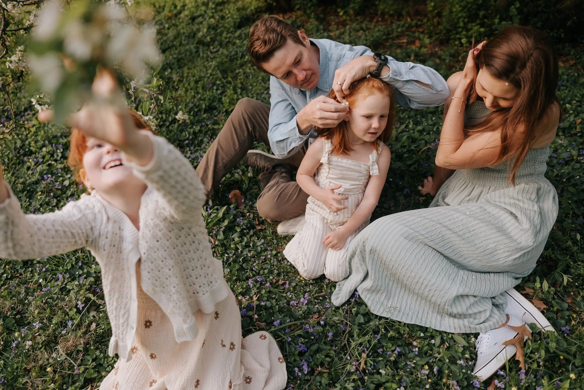 A family of five, including two adult women and three young girls, sitting and playing outside on a grassy area with small purple and white flowers, surrounded by green plants and trees.