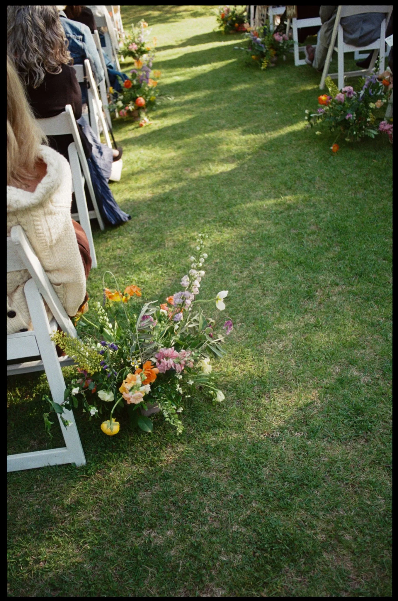 Outdoor wedding ceremony setup on grass with white chairs, colorful flower arrangements, and floral decorations along the aisle, under natural daylight.