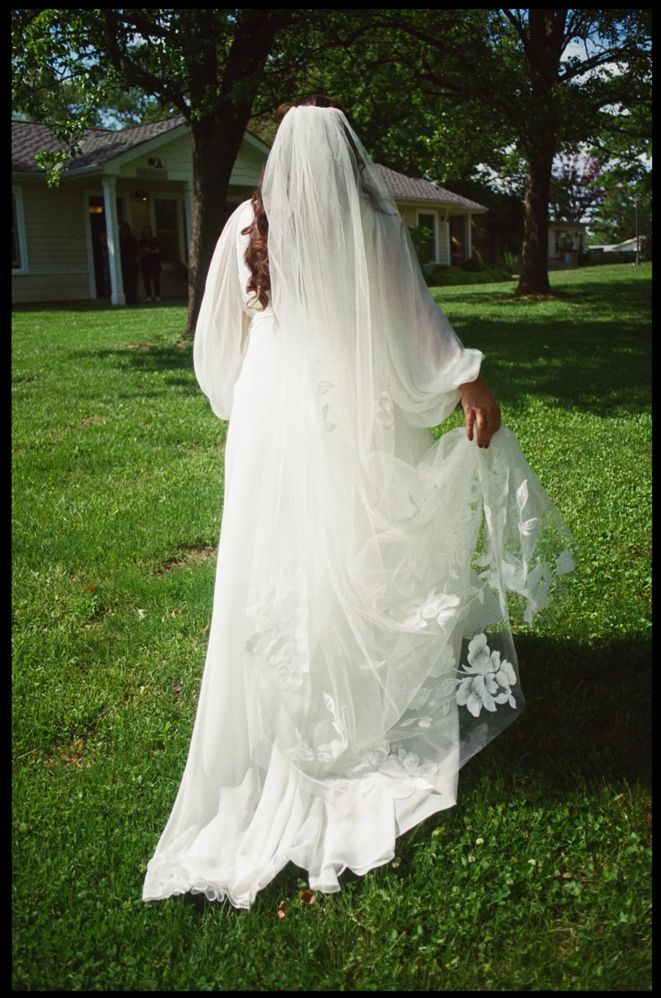 A bride wearing a white wedding dress with floral embroidery walking on green grass in a yard, with trees and a house in the background.