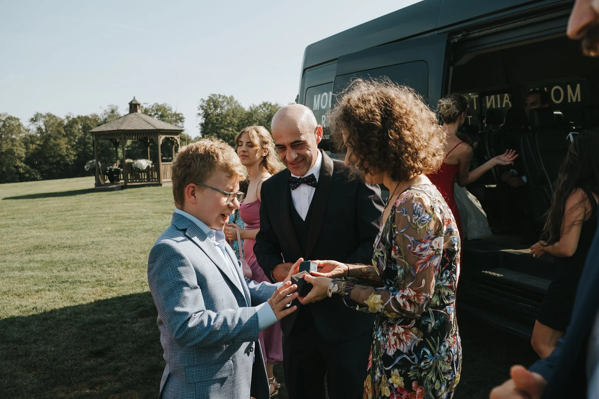A boy in a light blue checkered suit is receiving a ring in a small box from a woman with curly hair and a floral dress, while a man in a tuxedo and bow tie stands nearby, smiling. They are outdoors with a green lawn, trees, and a gazebo in the backg