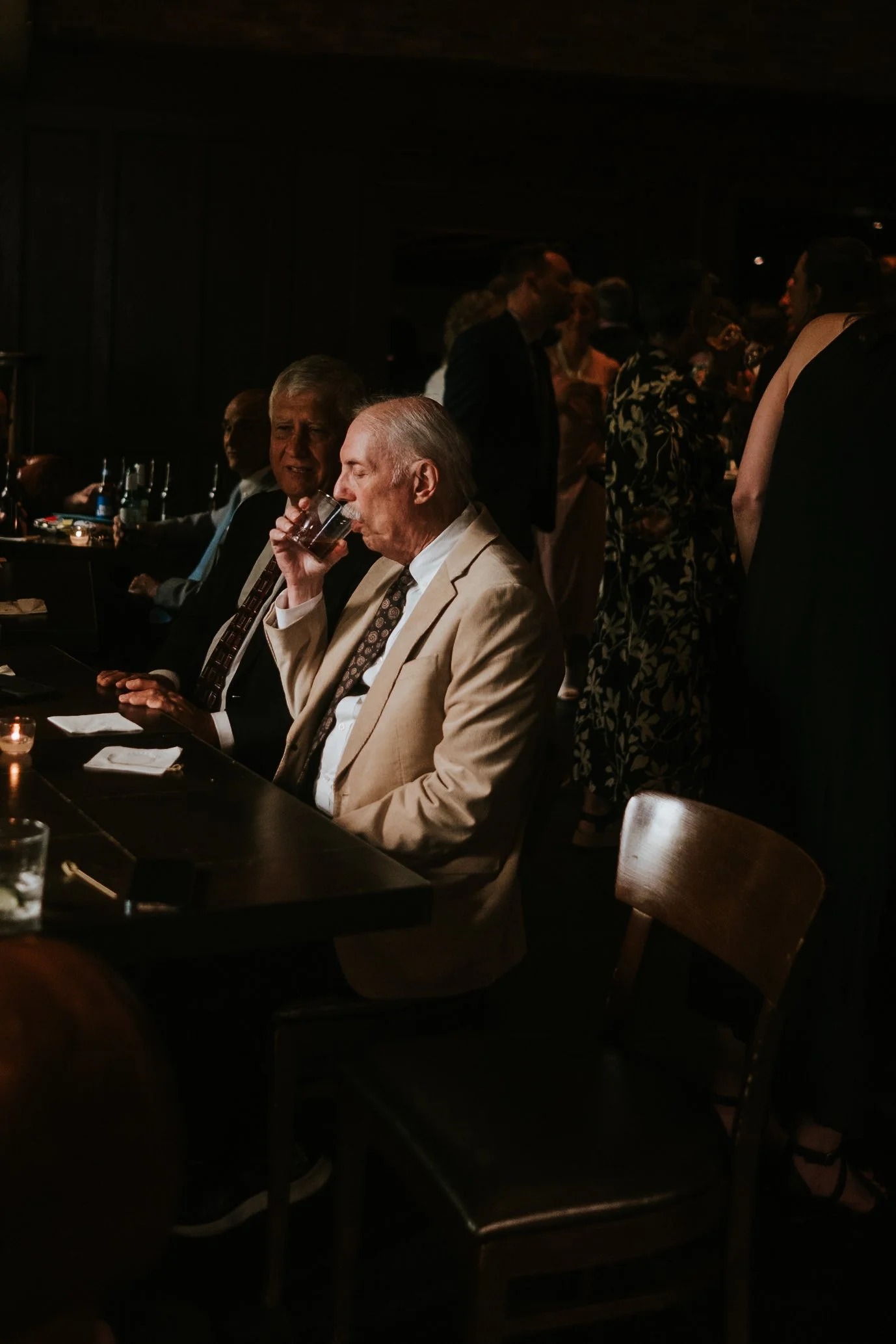 A dimly lit social gathering with mostly older adults, some sitting at a table and others standing and chatting in the background. The focus is on a man in a beige suit drinking from a glass, with other men seated nearby.