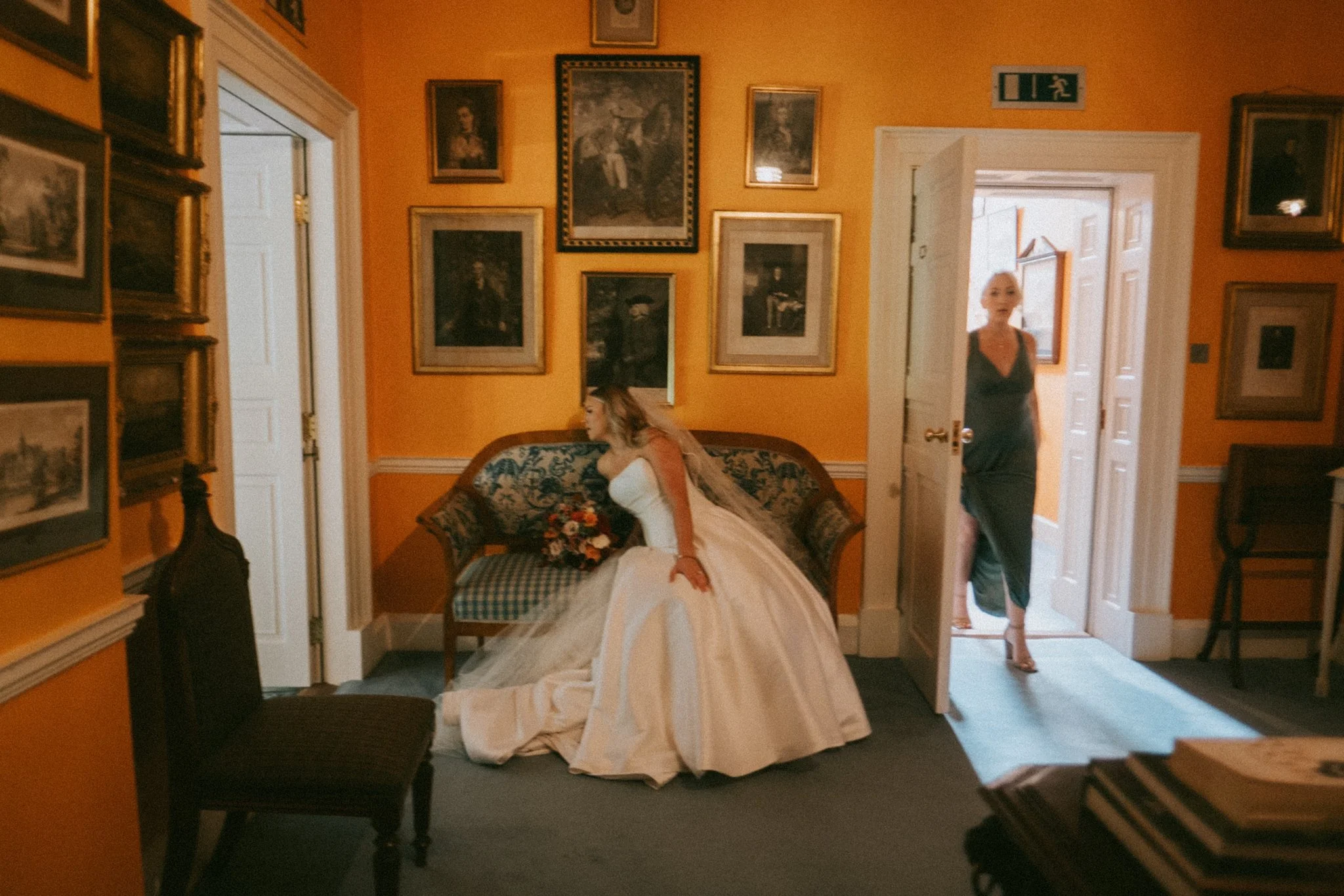 A woman in a wedding dress sitting on a vintage sofa with floral upholstery, holding a bouquet of flowers, in a room with orange walls decorated with framed paintings, and a woman in a black dress standing in the doorway.