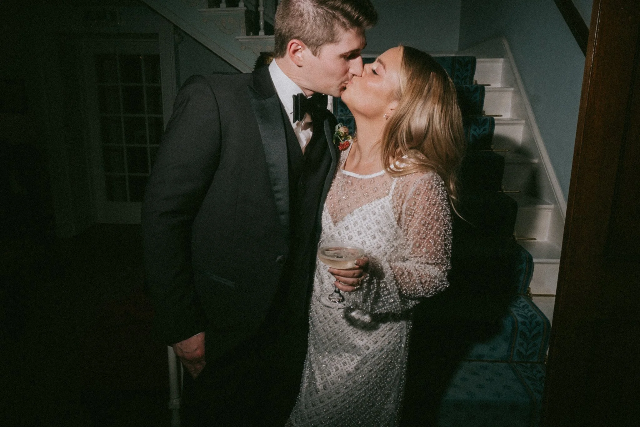 A couple is sharing a kiss at what appears to be a formal event. The man is wearing a tuxedo with a bow tie, and the woman is dressed in a sparkling, lace gown. She is holding a glass of champagne. They are standing next to a staircase in a dimly lit
