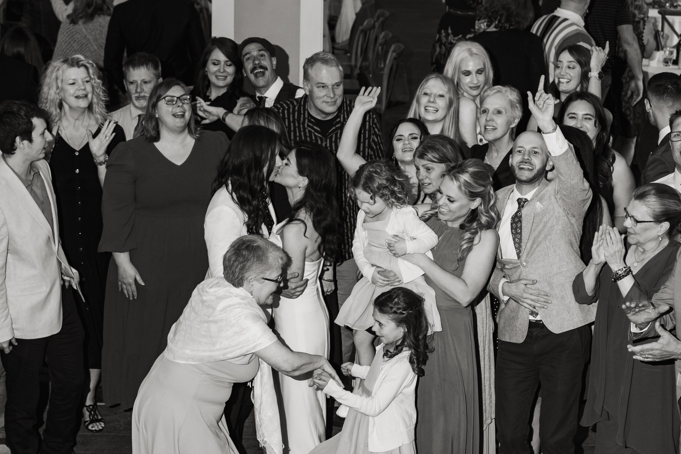 A large group of people gathered at a social event, with many smiling, chatting, and some raising their hands in the air. Two women are kissing in the foreground, while others are talking and enjoying themselves.