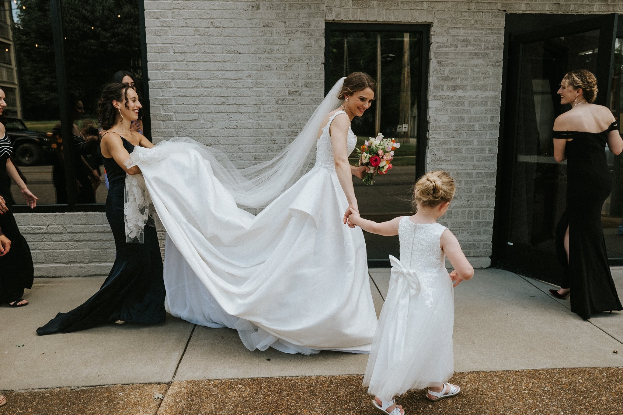 A bride in a white wedding dress holding a bouquet is walking outside on a sidewalk. She is surrounded by women in black dresses, some holding her dress to assist her. Two young girls in white dresses are also present, one holding the bride's hand. T