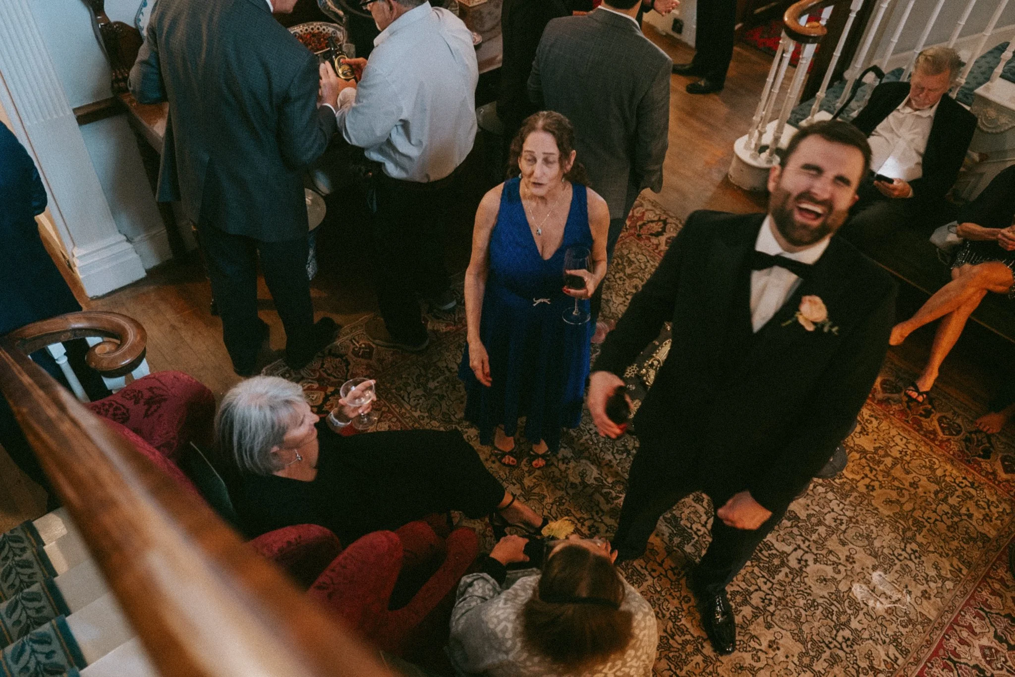 People dressed in formal attire enjoying a social gathering in a warmly lit room with a patterned carpet and wooden flooring.