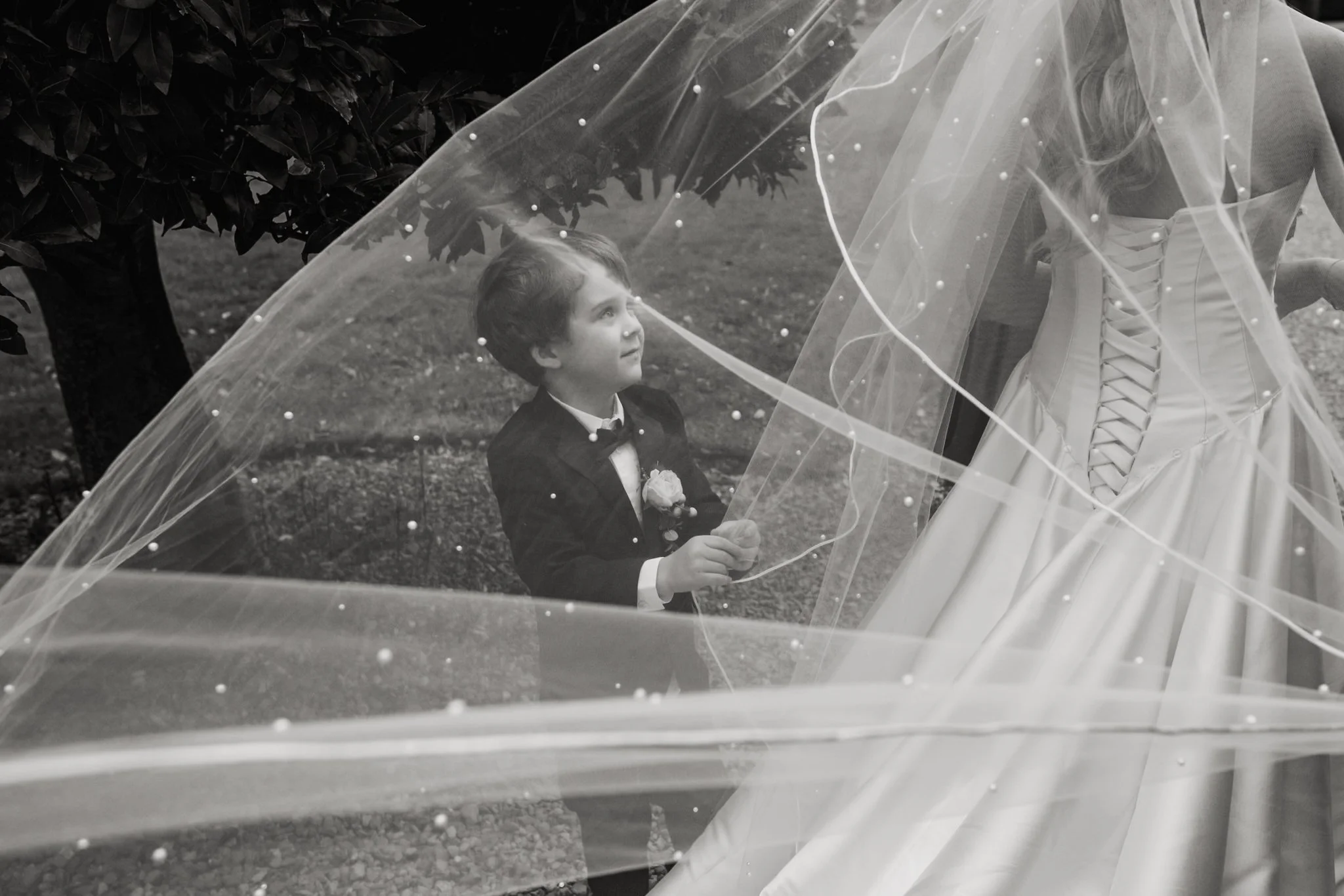 A young boy in a tuxedo looking at a woman in a wedding dress and veil under a tree.