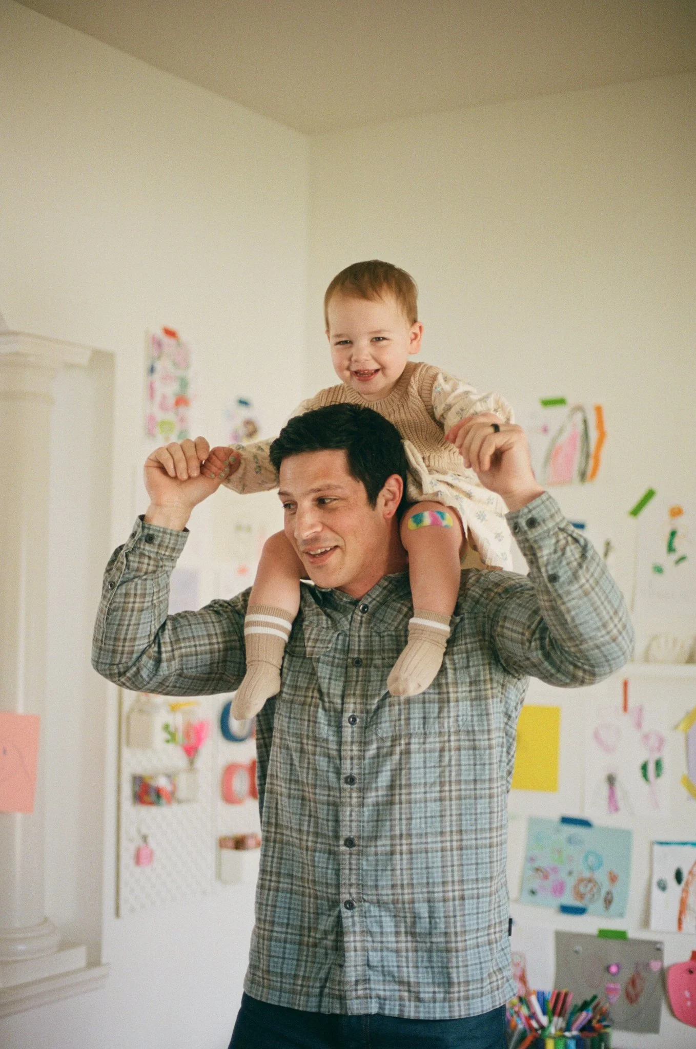 A man carrying a young child on his shoulders indoors. The child is smiling and holding the man's head. The background has colorful artwork and a white wall.
