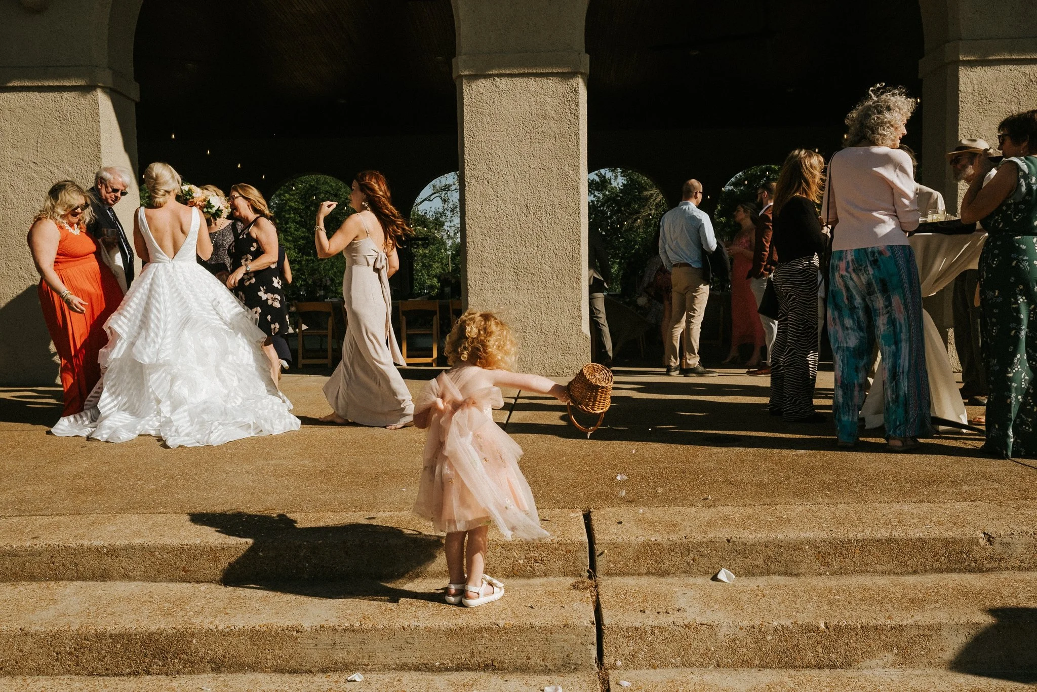 A group of people gathered under a bridge, with a woman in a white wedding dress and others dressed in various formal and casual attire, some mingling and some standing, during daytime. A small girl in a pink dress is in the foreground holding a litt