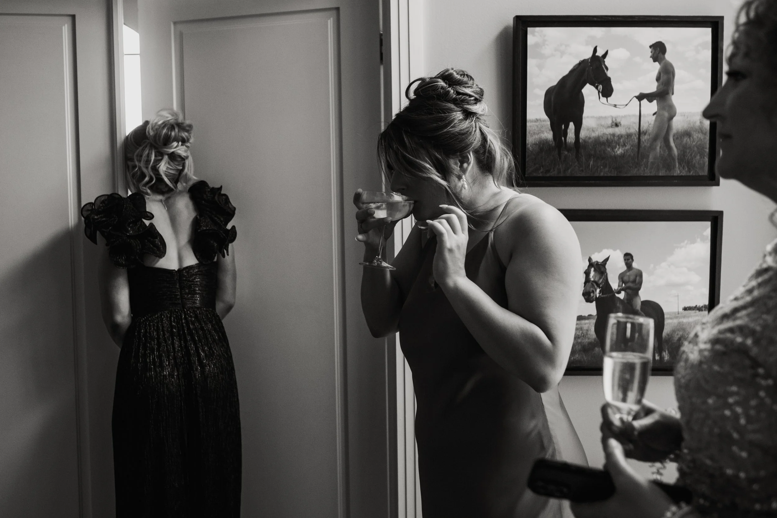 Black and white photo of a woman drinking from a cocktail glass at a social gathering, with a woman in a dress and another person partially visible, and framed equestrian photographs on the wall.