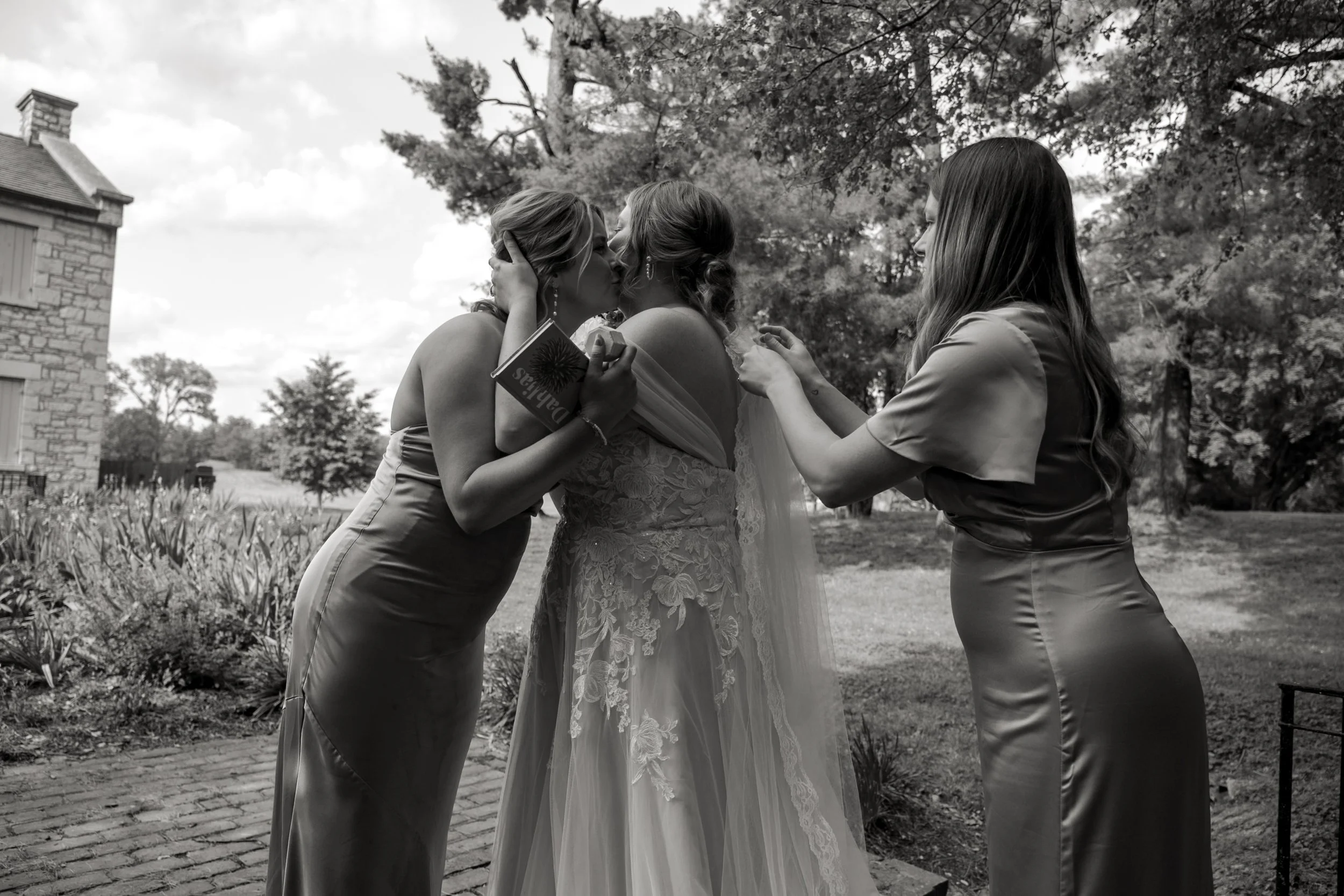 Three women in formal attire kissing and holding small objects outdoors on a sunny day.