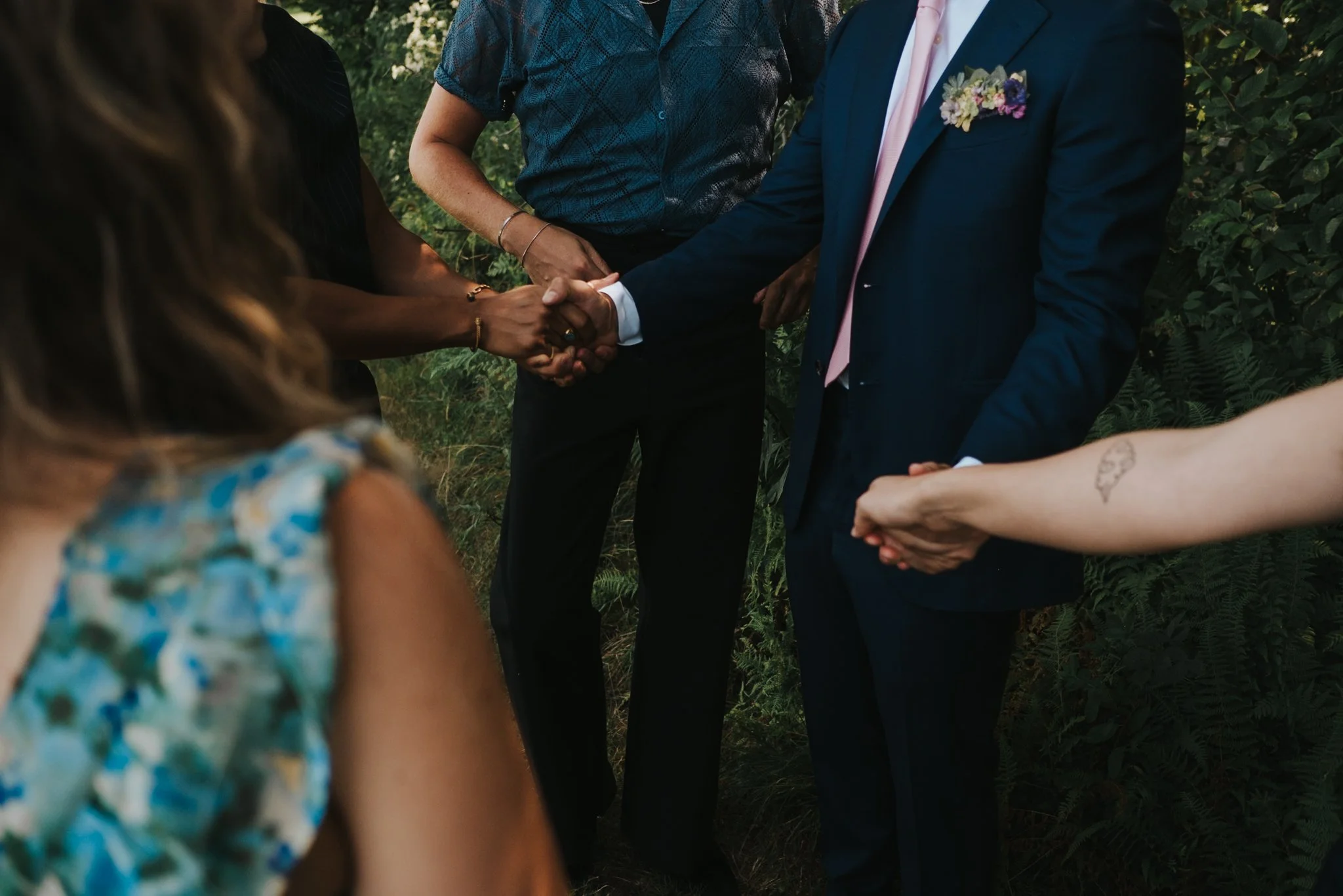 A group of people holding hands during an outdoor wedding or ceremony, standing among greenery.