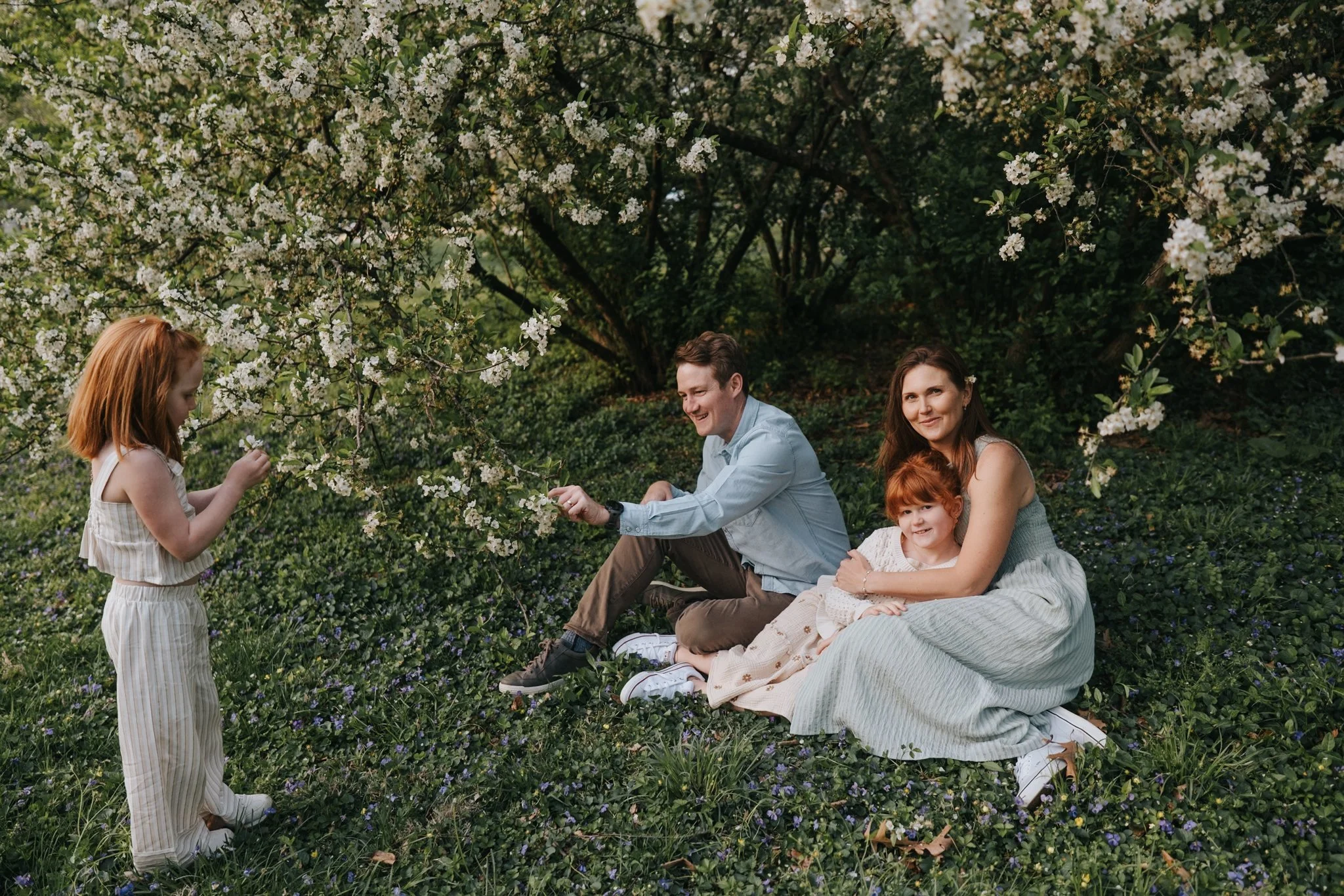 A family of four, including a mother, father, and two young girls, sitting and standing under a blooming tree in a lush green garden during daytime.
