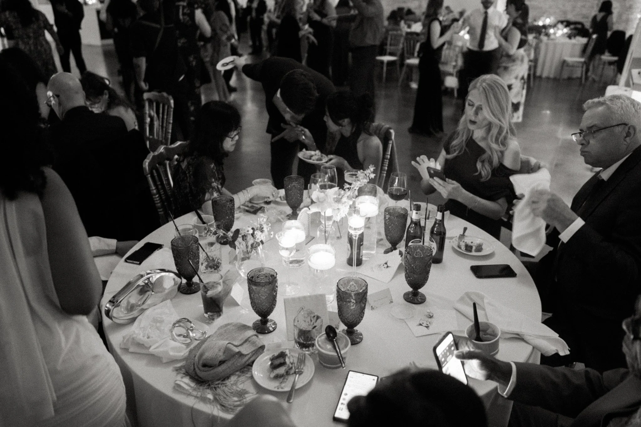 A black and white photo of a dinner event with many people seated around tables, some engaged with their phones, others talking, with a lively, social atmosphere.