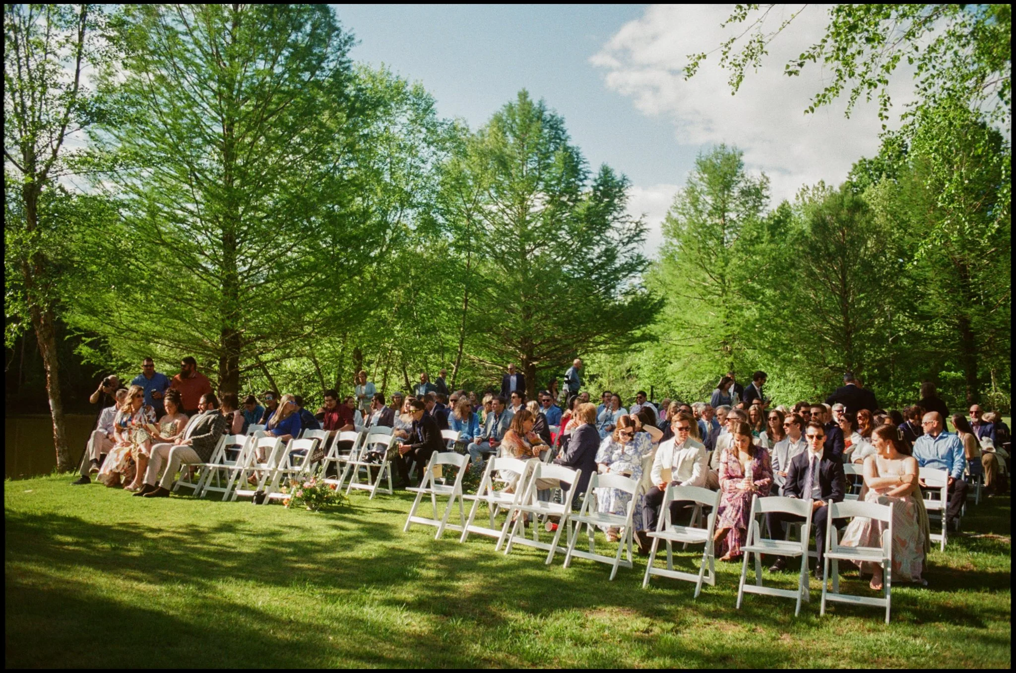 A gathering of people seated on white chairs outdoors in a green park, surrounded by tall trees and blue sky.